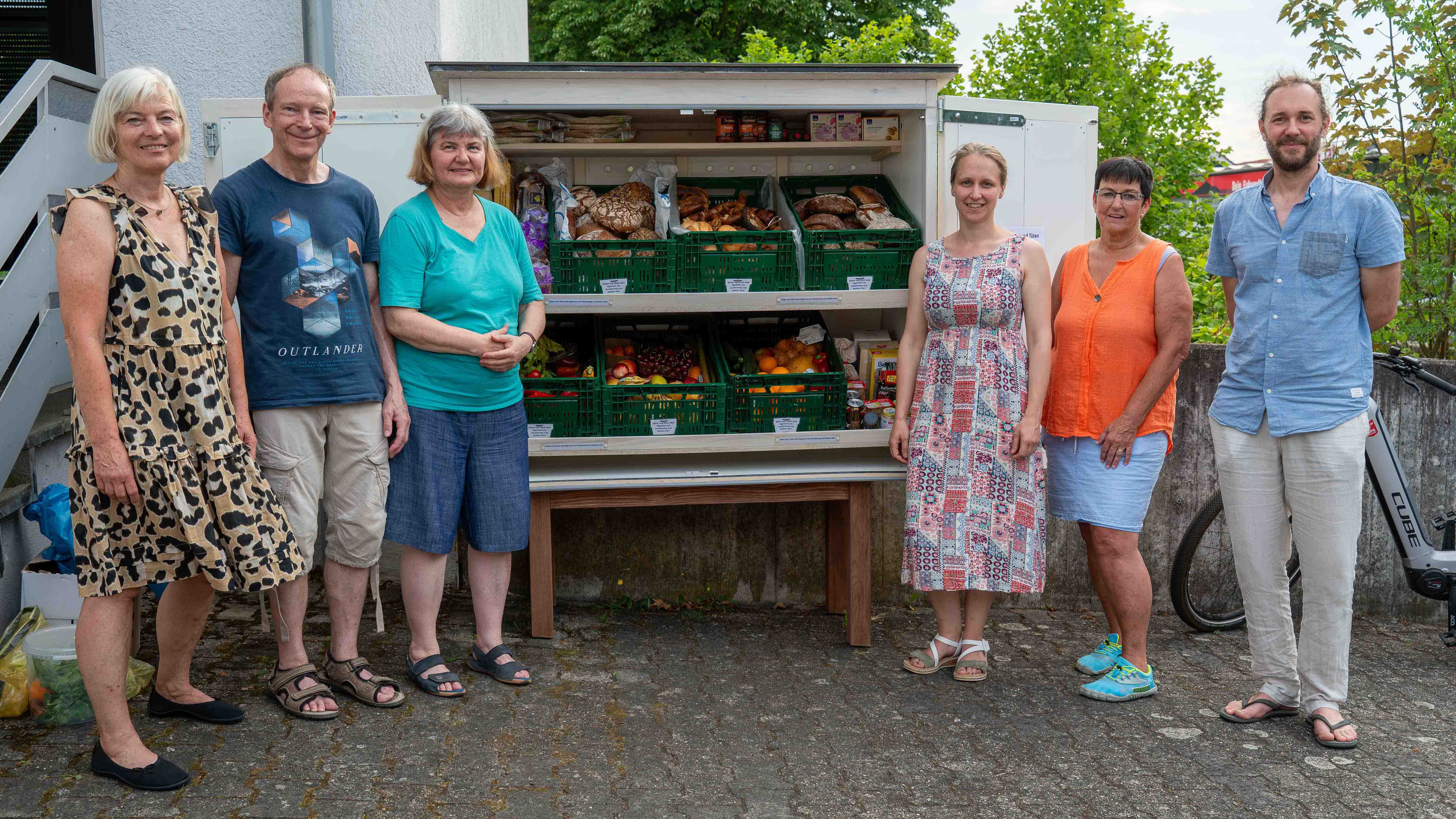 Zwingenberg_Fairteiler_C_Michael_Ränker_1_16zu9.jpg Sechs Personen stehen vor einem öffentlichen Lebensmittel-Fairteiler mit Brot, Obst und Gemüse und präsentieren gemeinsam das Angebot.