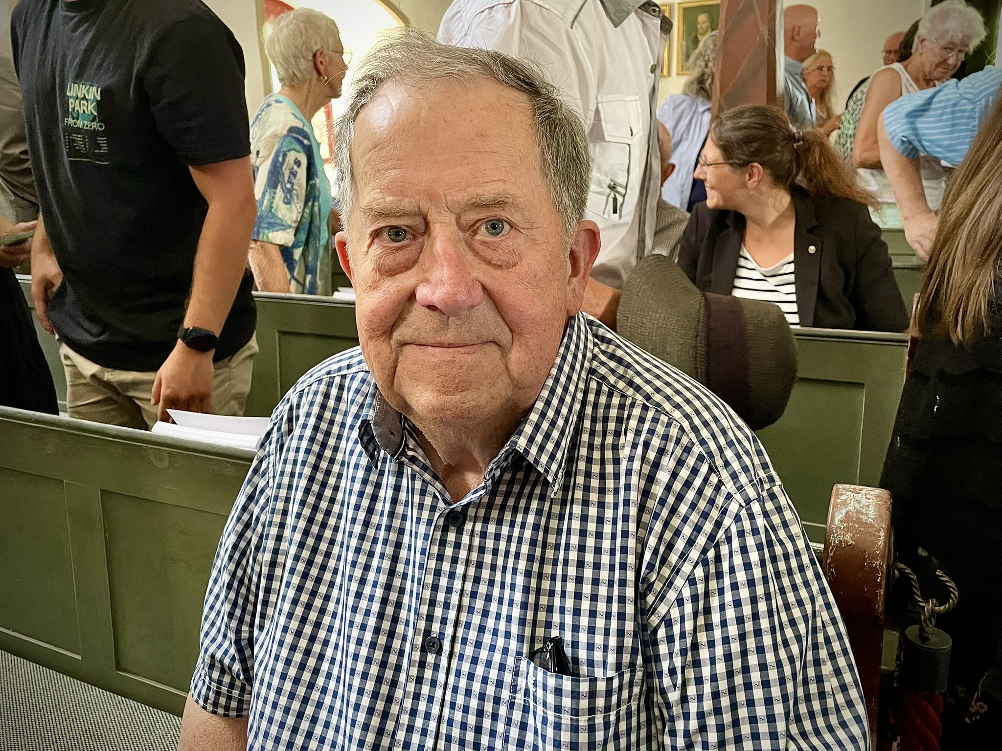 Otto Peter sitzt lächelnd in einer Kirchenbank und blickt in die Kamera. Der 83-Jährige trägt ein blau-weiß kariertes Hemd. Im Hintergrund sind weitere Gottesdienstbesucherinnen und -besucher in der Groß-Eichener Kirche zu sehen.