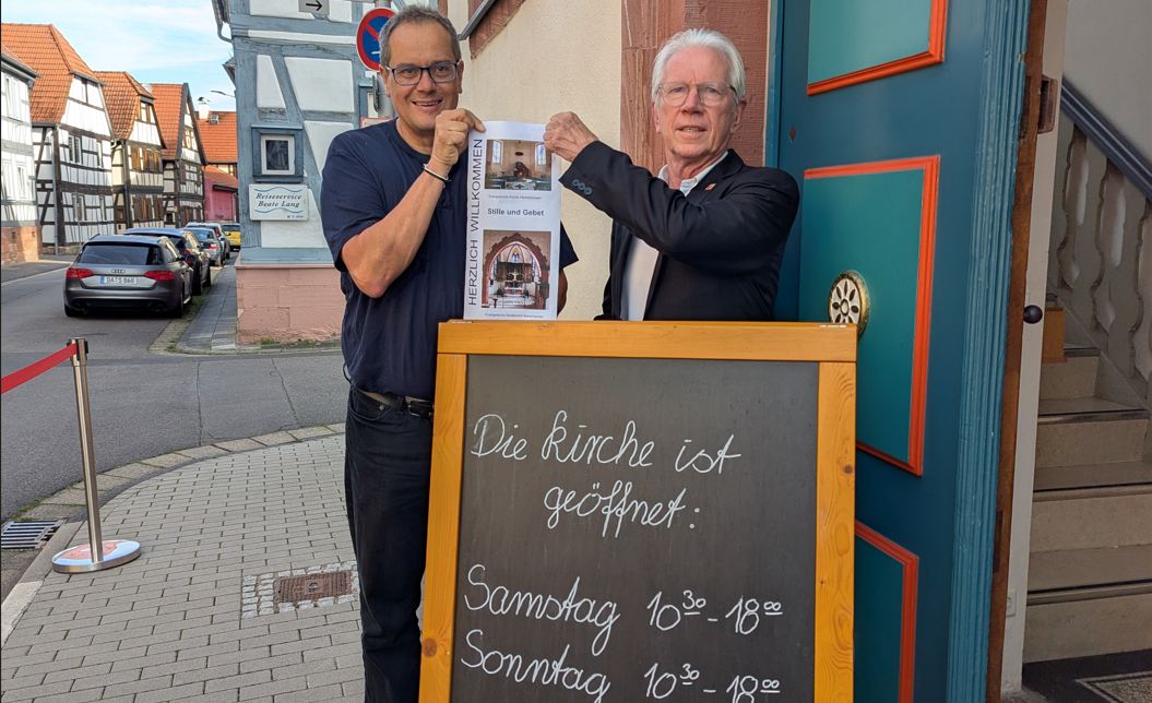 2 Personen stehen vor einem Schild und präsentieren einen Flyer.