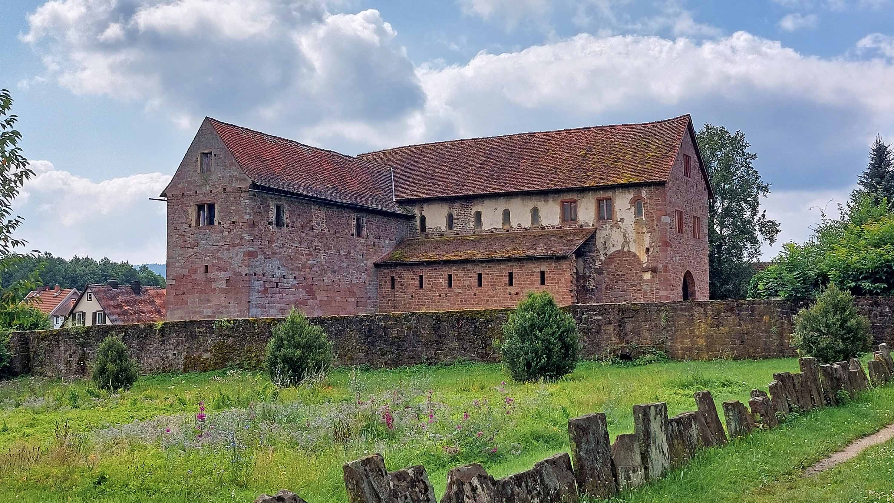 Historische steinerne Basilika mit roten Ziegeldächern hinter einer niedrigen Mauer, davor eine Wiese mit Sträuchern unter bewölktem Himmel.