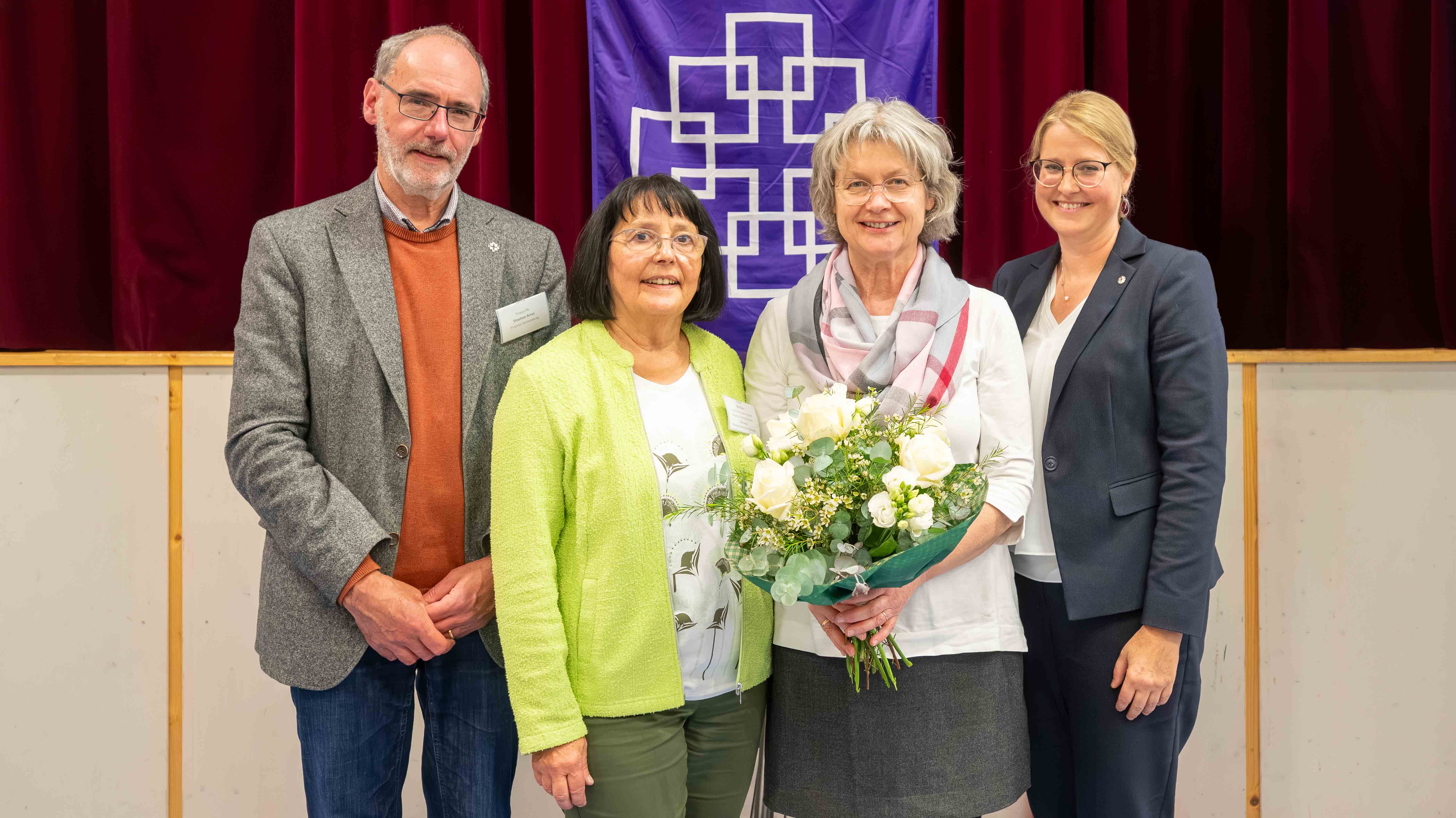 Herbstsynode_2026_C_Michael_Ränker_29_16zu9.jpg Vier Personen stehen bei der Herbstsynode des Evangelischen Dekanats Bergstraße nebeneinander vor einem roten Bühnenvorhang und einem violetten Banner mit weißem kirchlichem Symbol. Von links: Propst Stephan Arras im grauen Sakko, Präses Ute Gölz in hellgrüner Jacke, die wiedergewählte stellvertretende Dekanin Silke Bienhaus mit einem großen weiß-grünen Blumenstrauß sowie Dekanin Sonja Mattes im dunkelblauen Hosenanzug. Alle vier lächeln in die Kamera.