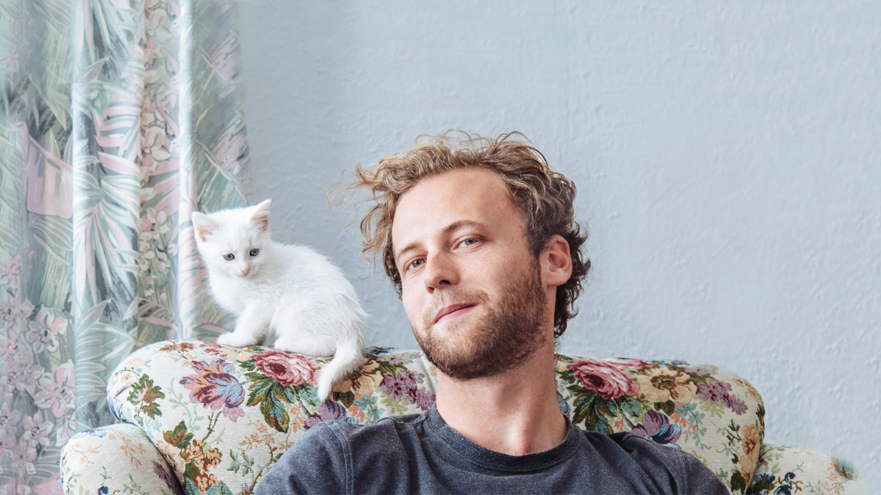 Junger Mann sitzt auf einem Sessel mit Blumenmuster, auf der Lehne sitzt eine junge, weiße Katze
