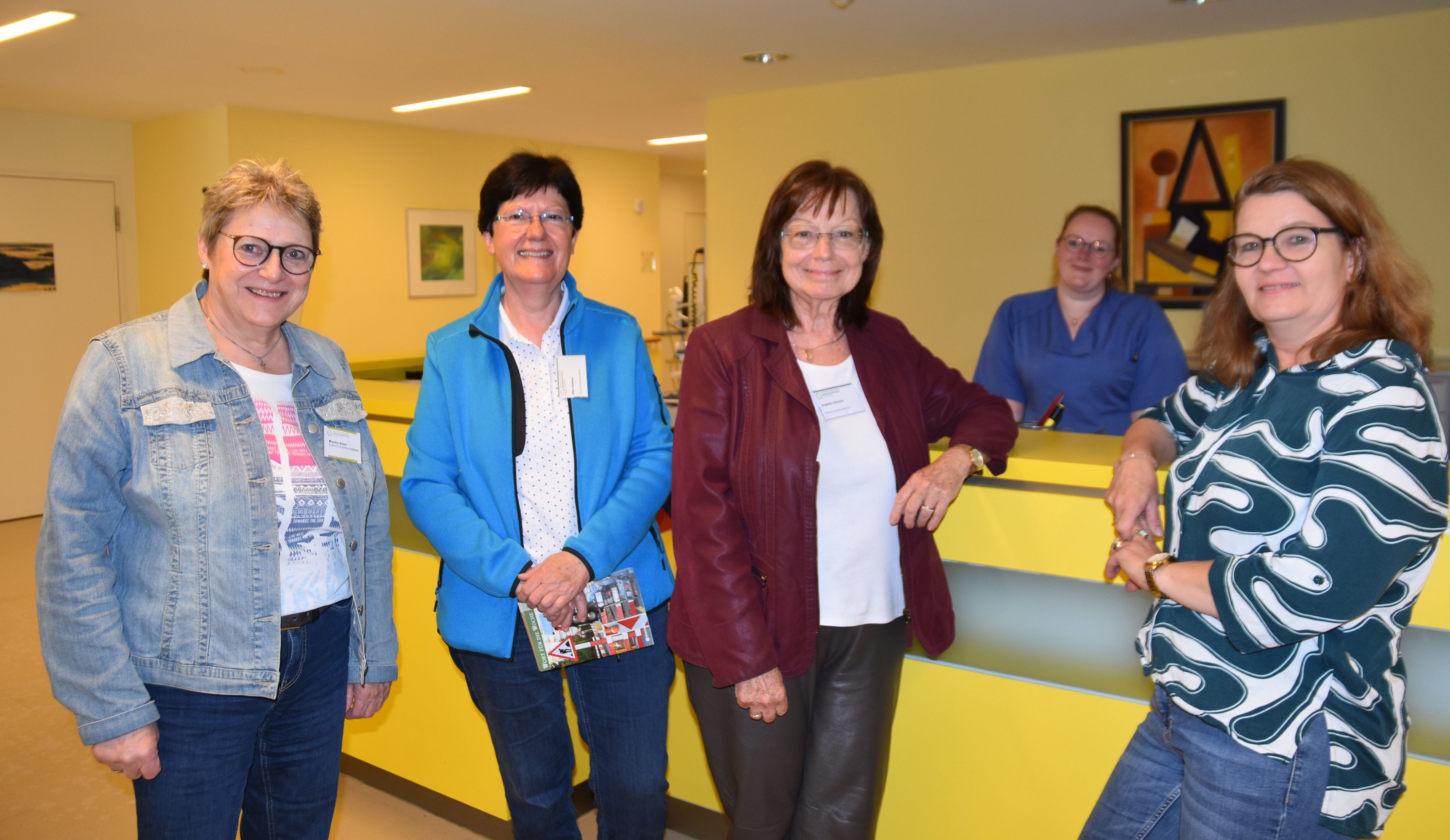 Vier Frauen in Alltagskleidung stehen an einem Tresen auf einem Krankenhausflur und blicken in die Kamera. Hinter dem Tresen steht eine Krankenschwester.