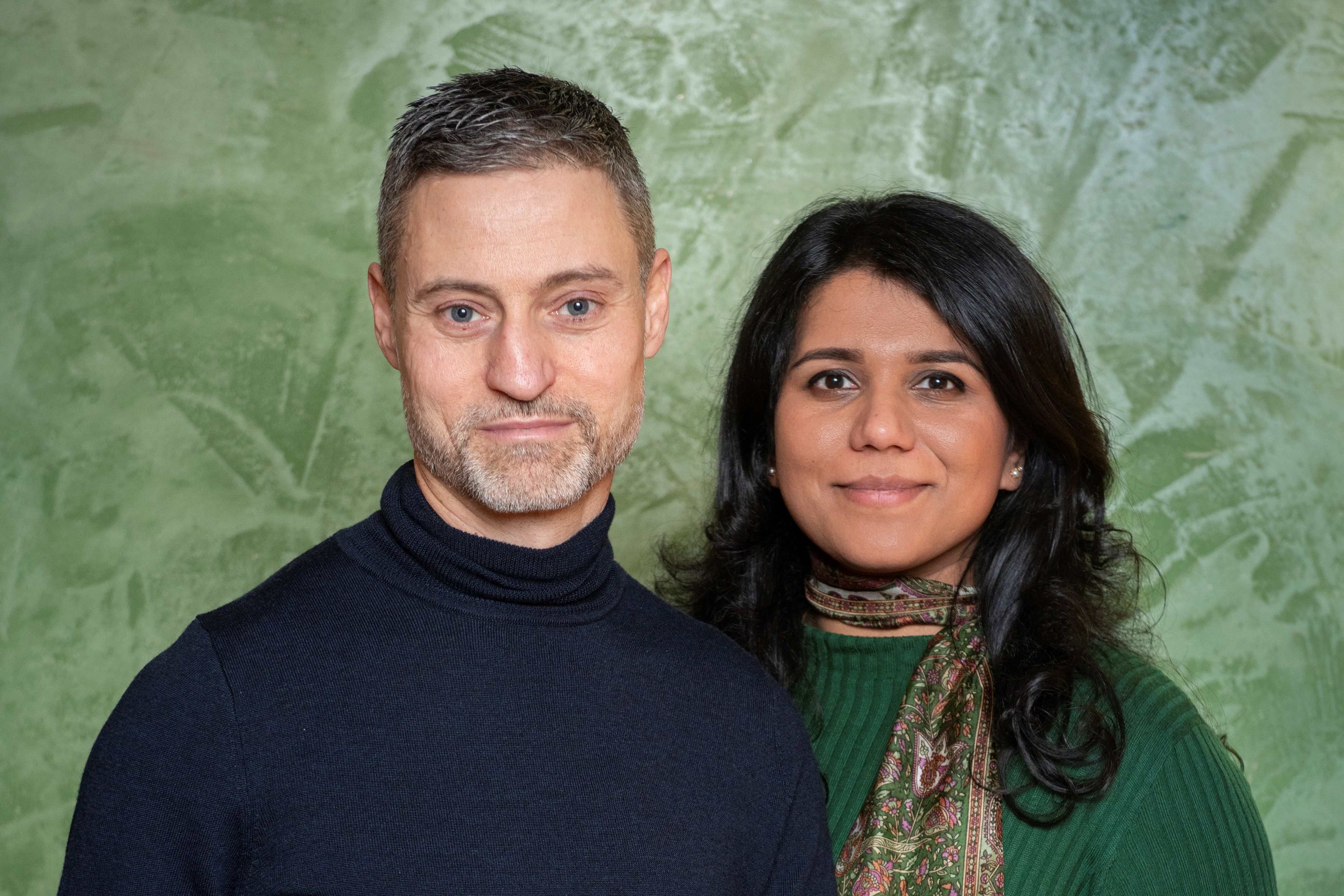 Porträt Meron Mendel und Saba-Nur Cheema vor einem grünen Hintergrund. Er trägt eine dunkelblauen Pullover, sie einen grünen und ein buntes Halstuch.