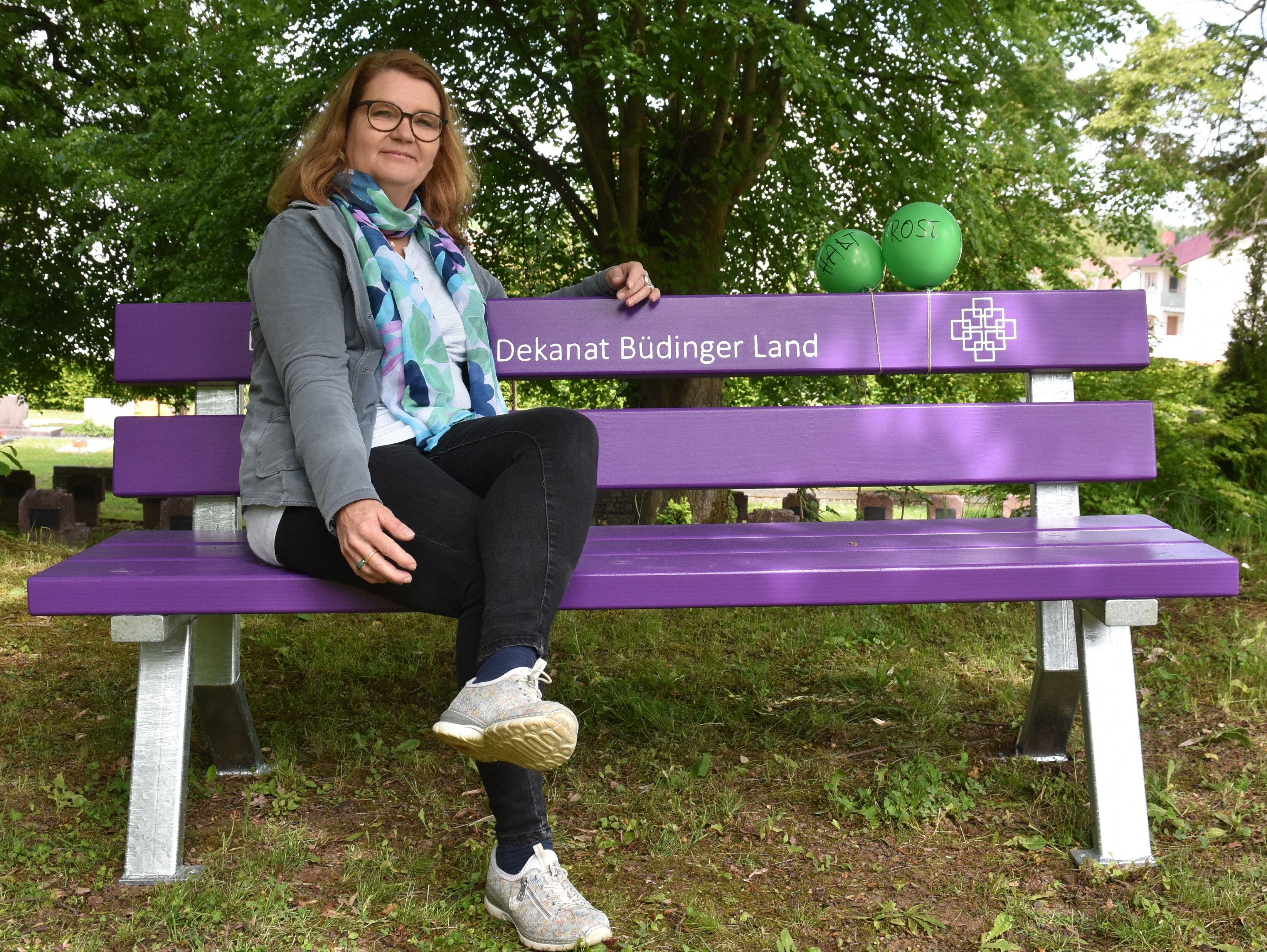 Astrid Pfarschner hat in entspannter Haltung auf der Trauerbank Platz genommen. Die Bank iägt die Aufschrift ec. Dekanat Büdinger Land in weiß sowie zwei Facettenkreuze. Im Hintergrund sieht man Bäume
