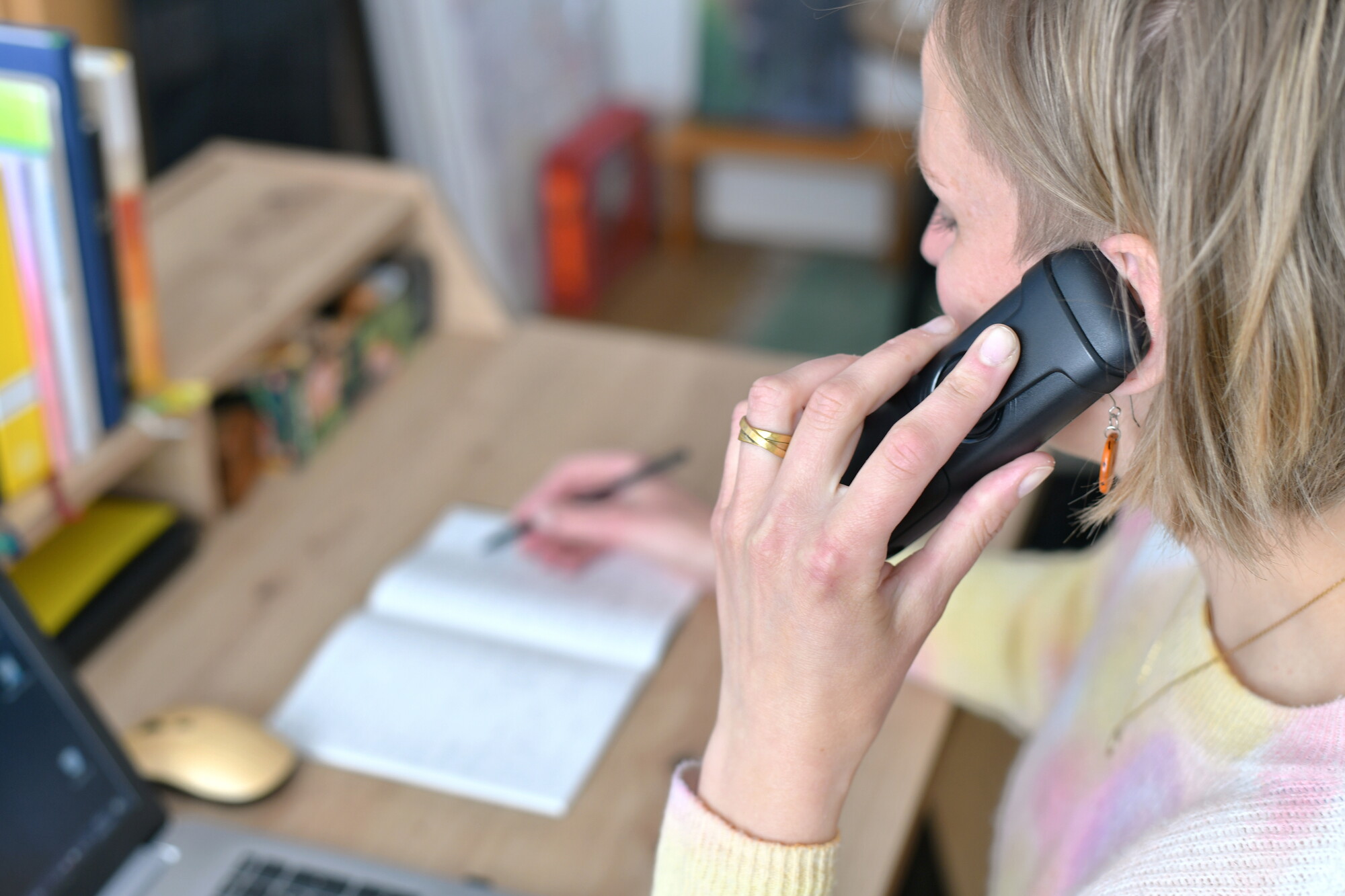 Pfarrerin Nele Schomakers in einem Büro beim Telefonieren - seitlich aufgenommen mit Blick auf den Bürotisch.  In der linken Hand das Telefon, in der rechten Hand ein Kugelschreiber um Notizen zu machen. Im Vordergrund die Hand und der Telefonhörer am Ohr.