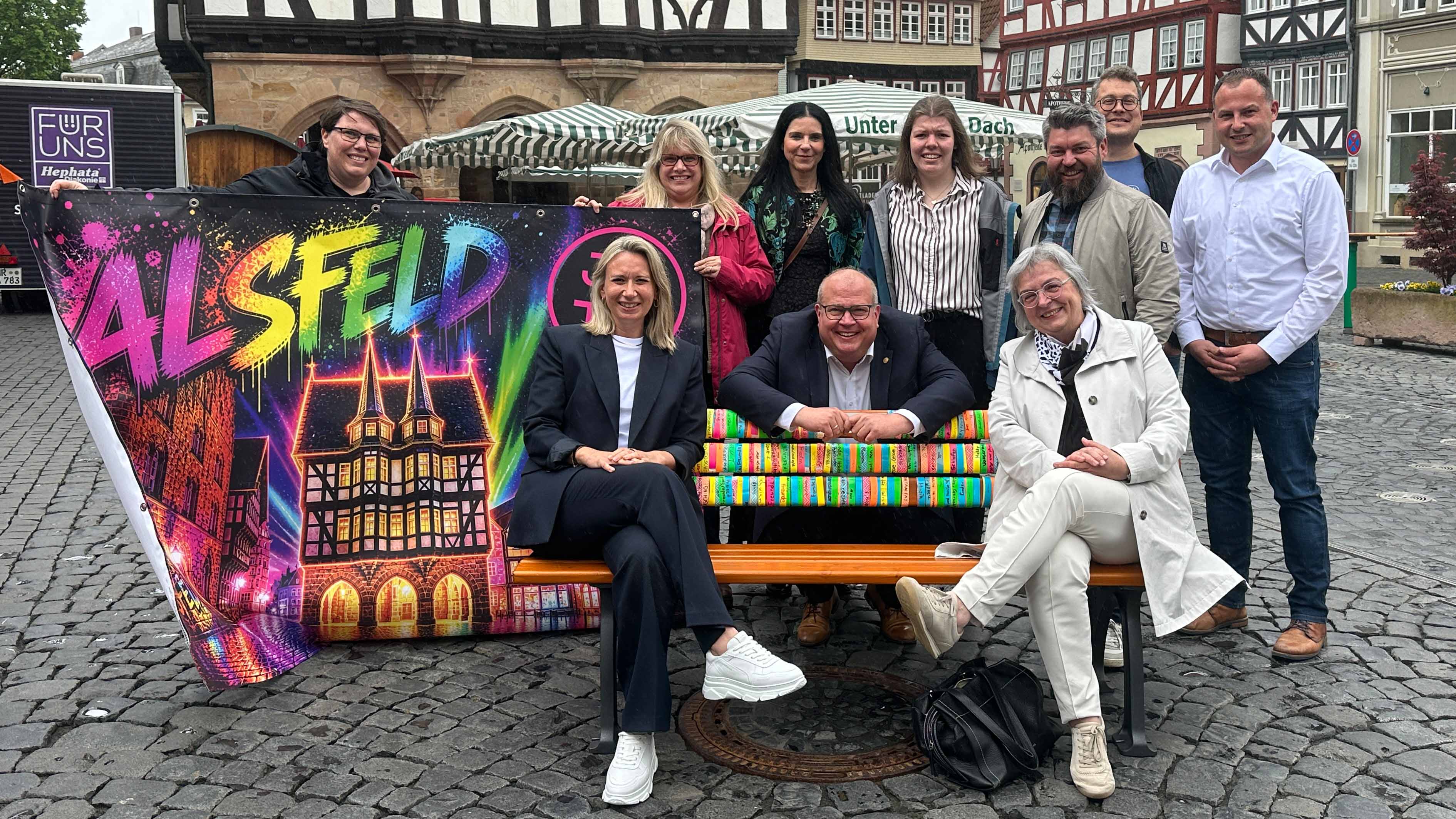 Gruppe von zehn Personen auf dem Marktplatz in Alsfeld hinter einer bunt gestalteten Bank und einem Banner ift „Amit der Aufschrlsfeld“.