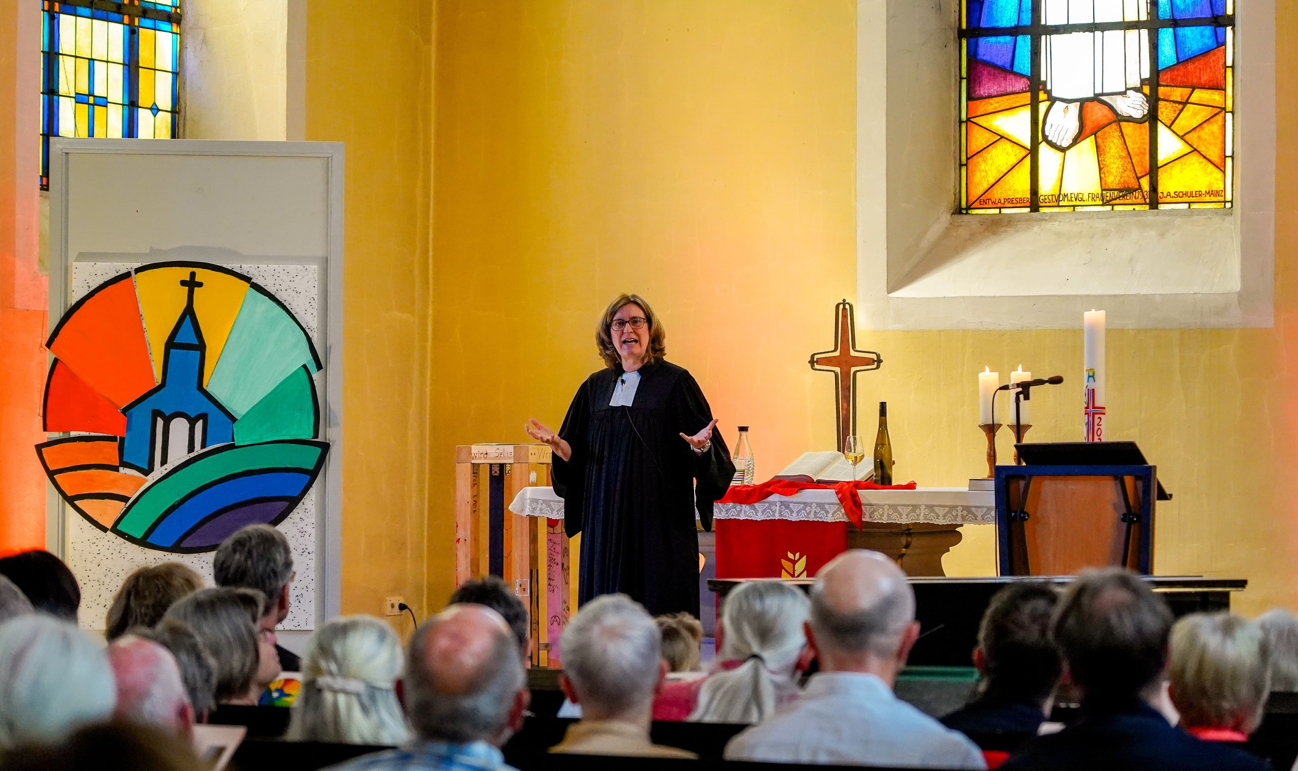 Eine Person in schwarzer Robe hält eine Predigt vor einer Gemeinde in einer Kirche mit bunten Glasfenstern und einem farbenfrohen Wandgemälde.
