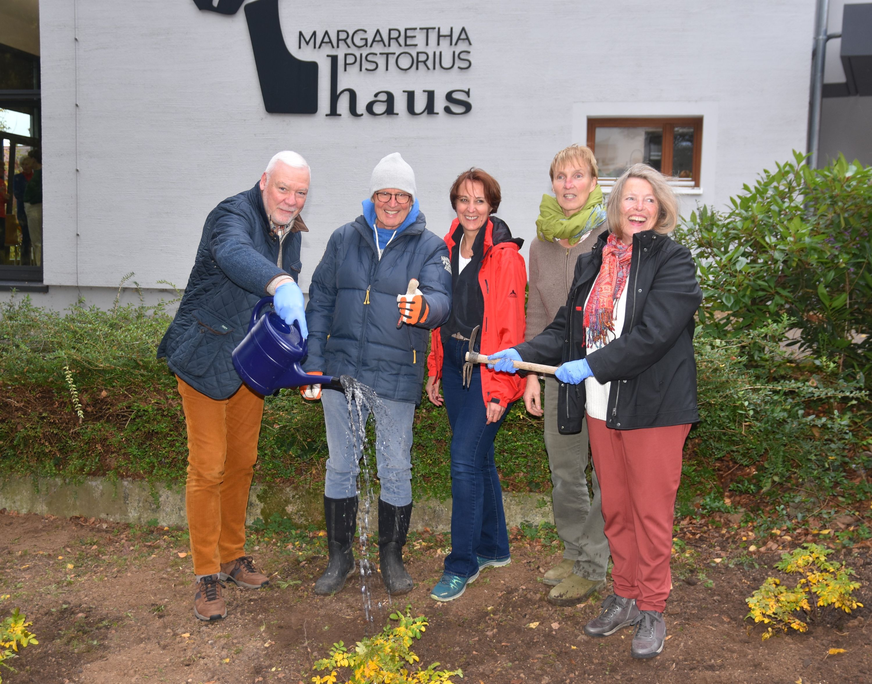 Zwei Männer, drei Frauen stehen im Johanniterpark auf Erdboden und blicken in die Kamera. Im Boden erkennt man die Rosenpflanzen. Die Menschen halten Gießkanne und Gartenschere und tragen Funktions- bzw. Arbeitskleidung. Entspannte Runde. Im Hintergrund sieht man das Margaretha-Pistorius-Haus.