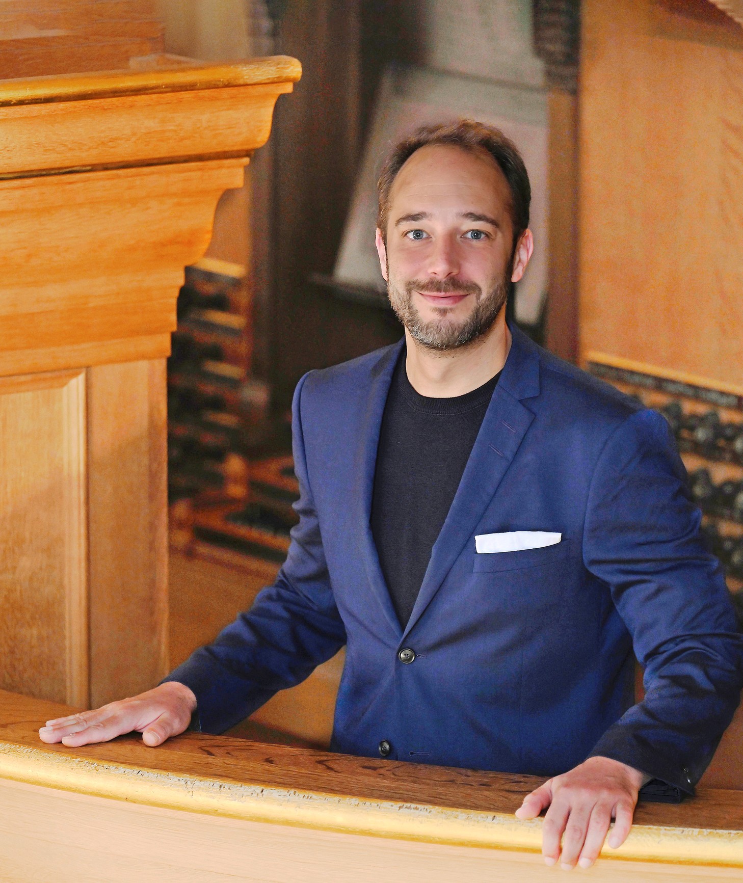 Ein Mann mit kurzem Bart trägt ein dunkelblaues Sakko über einem schwarzen Shirt und steht mit beiden Händen an einer hölzernen Brüstung im Innenraum einer Kirche, im Hintergrund eine Orgel.
