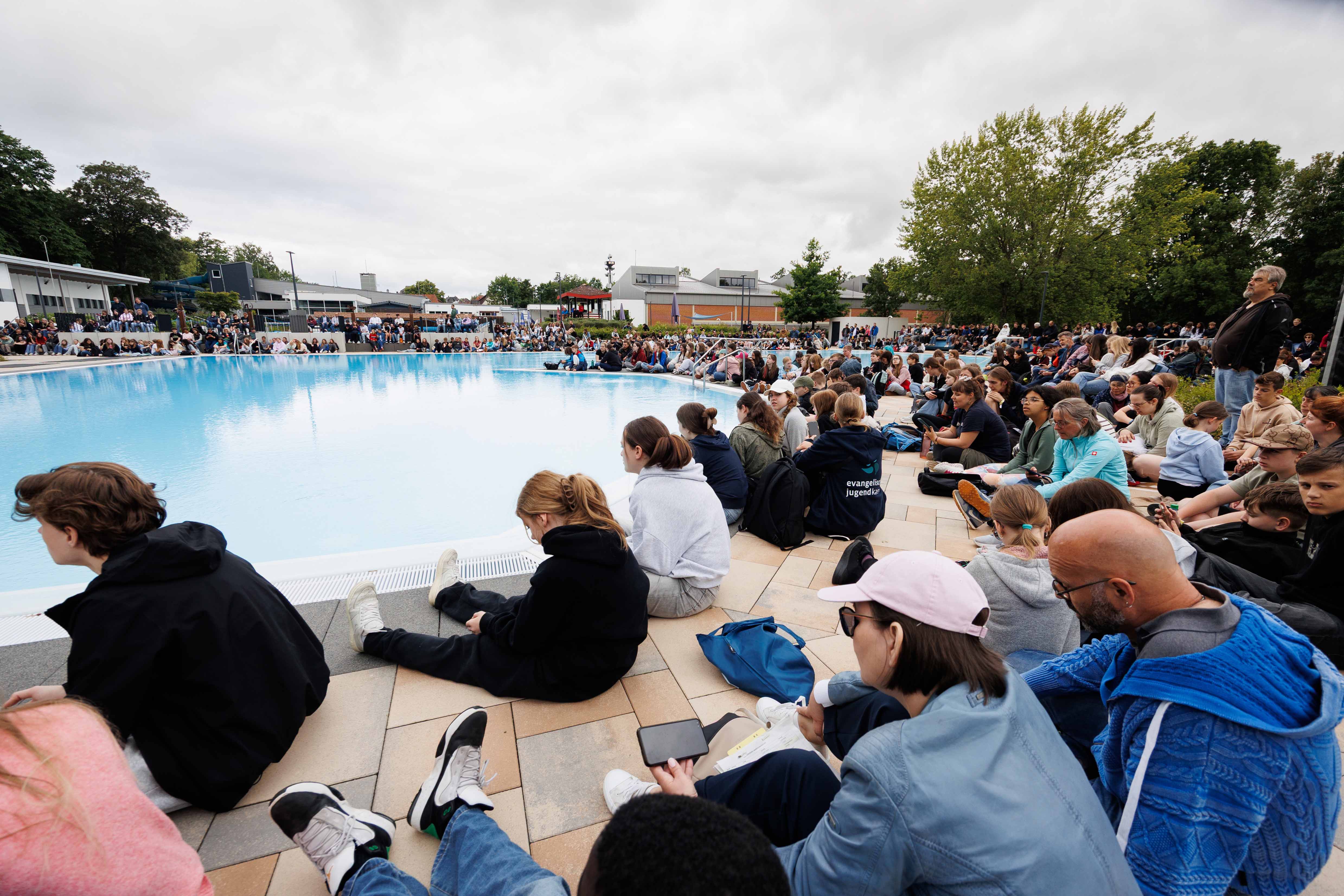 05-06-JKT-Freitag-Schwimmbadgottesdienst - Peter-Bongard (1) Kopie.jpg Ein großes Freibadbecken steht im Mittelpunkt. Entlang des gesamten Beckenrandes sitzen zahlreiche Menschen unterschiedlichen Alters auf den Fliesen oder auf den Tribünenbereichen. Im Hintergrund sind Gebäude des Freibads sowie Bäume zu sehen. Der Himmel ist stark bewölkt. Die Aufmerksamkeit der Anwesenden richtet sich auf einen Bereich am gegenüberliegenden Beckenrand, wo eine Veranstaltung stattfindet.