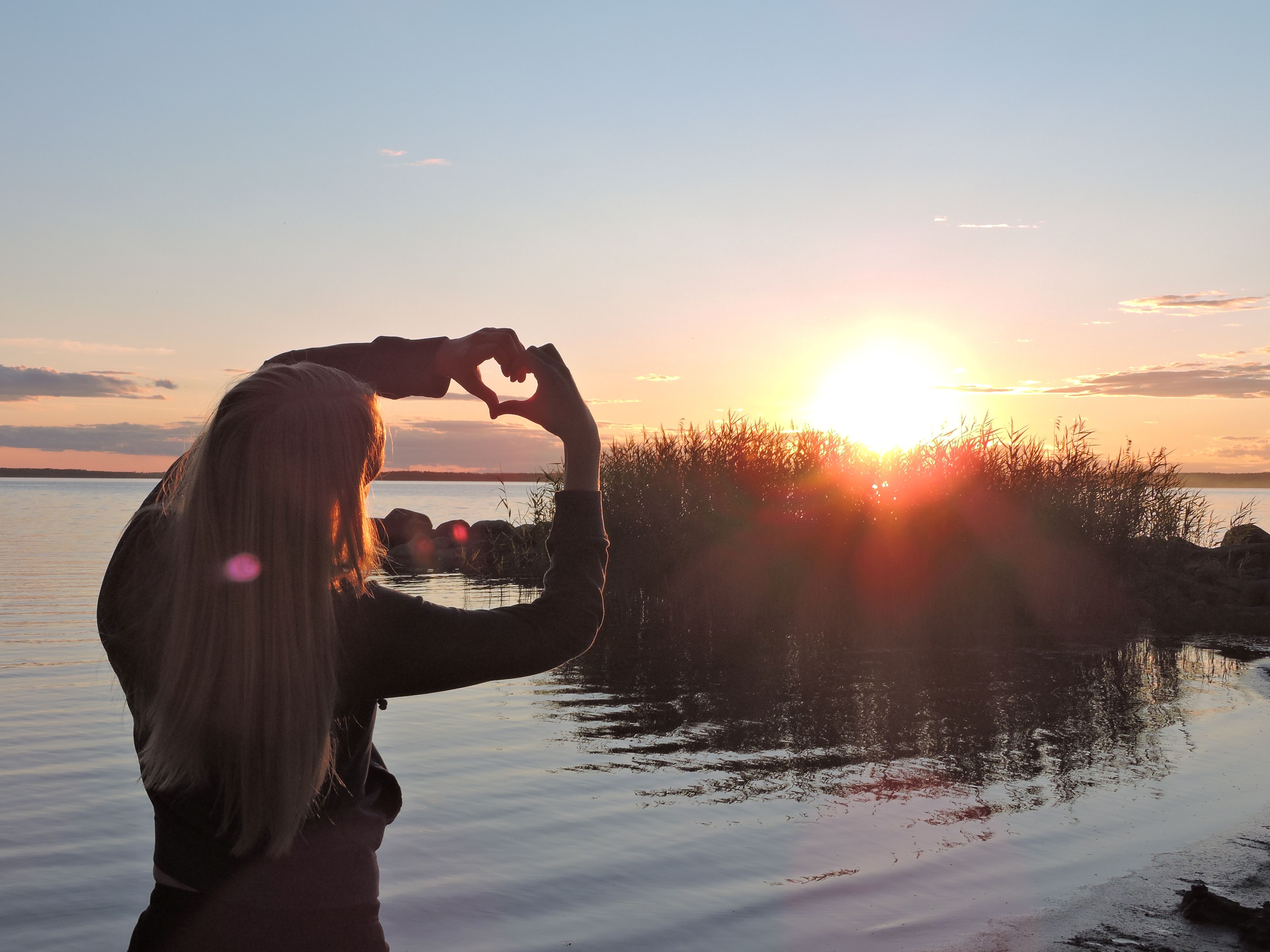 Eine Jugendliche steht bei Sonnenuntergang am Seeufer in Schwden und formt mit ihren Händen ein Herz