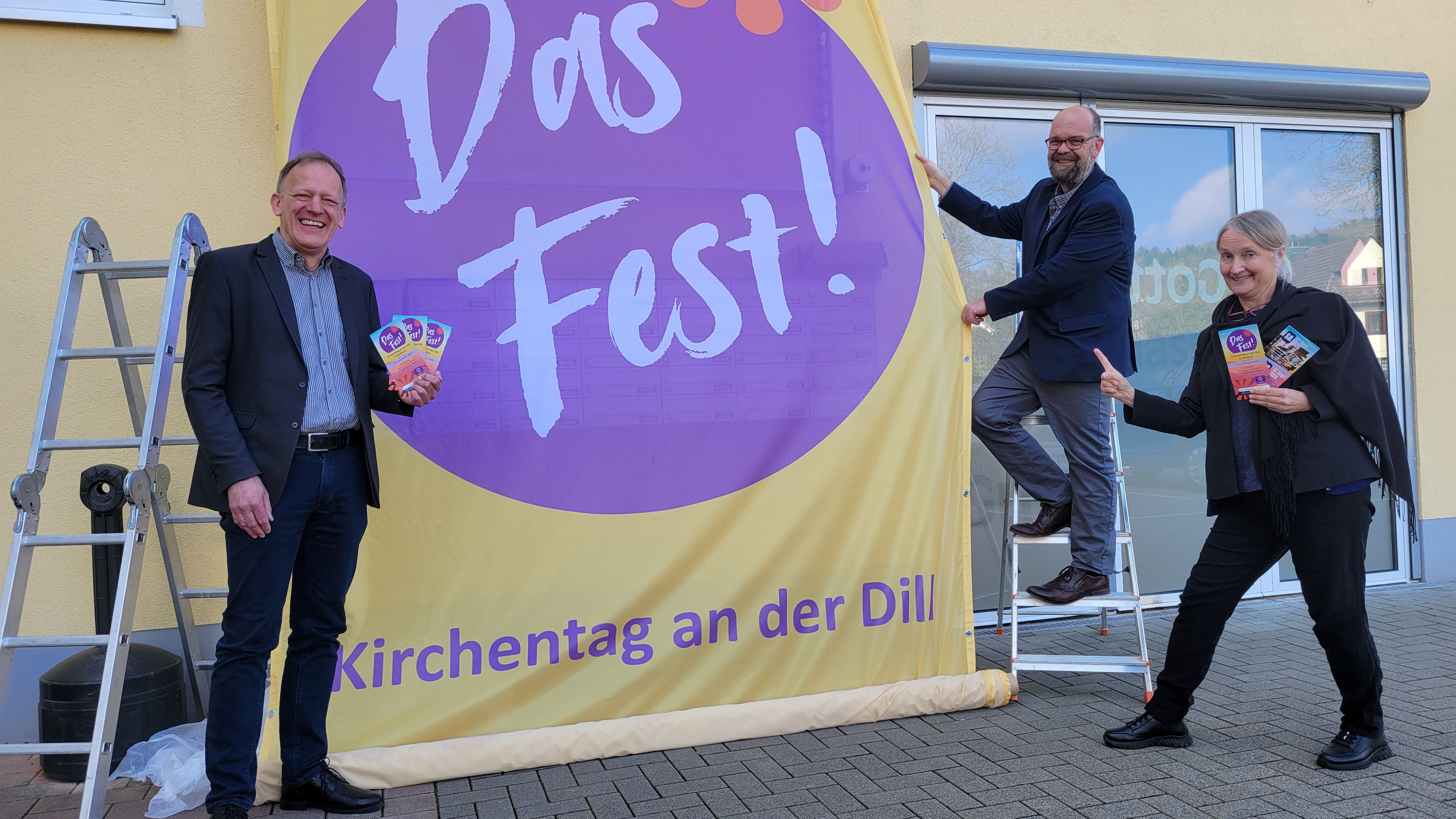 Mit dem Super-Banner „Das Fest!“ wirbt das Evangelische Dekanat an der Dill am Herborner „Haus der Kirche und Diakonie“ für den Dill-Kirchentag am 12. Und 13. September 2026. Präses Dr. Wolfgang Wörner (von links), Dekan Andree Best und die stellvertretende Dekanin Anja Vollendorf freuen sich über den Austausch und die Begegnung.
