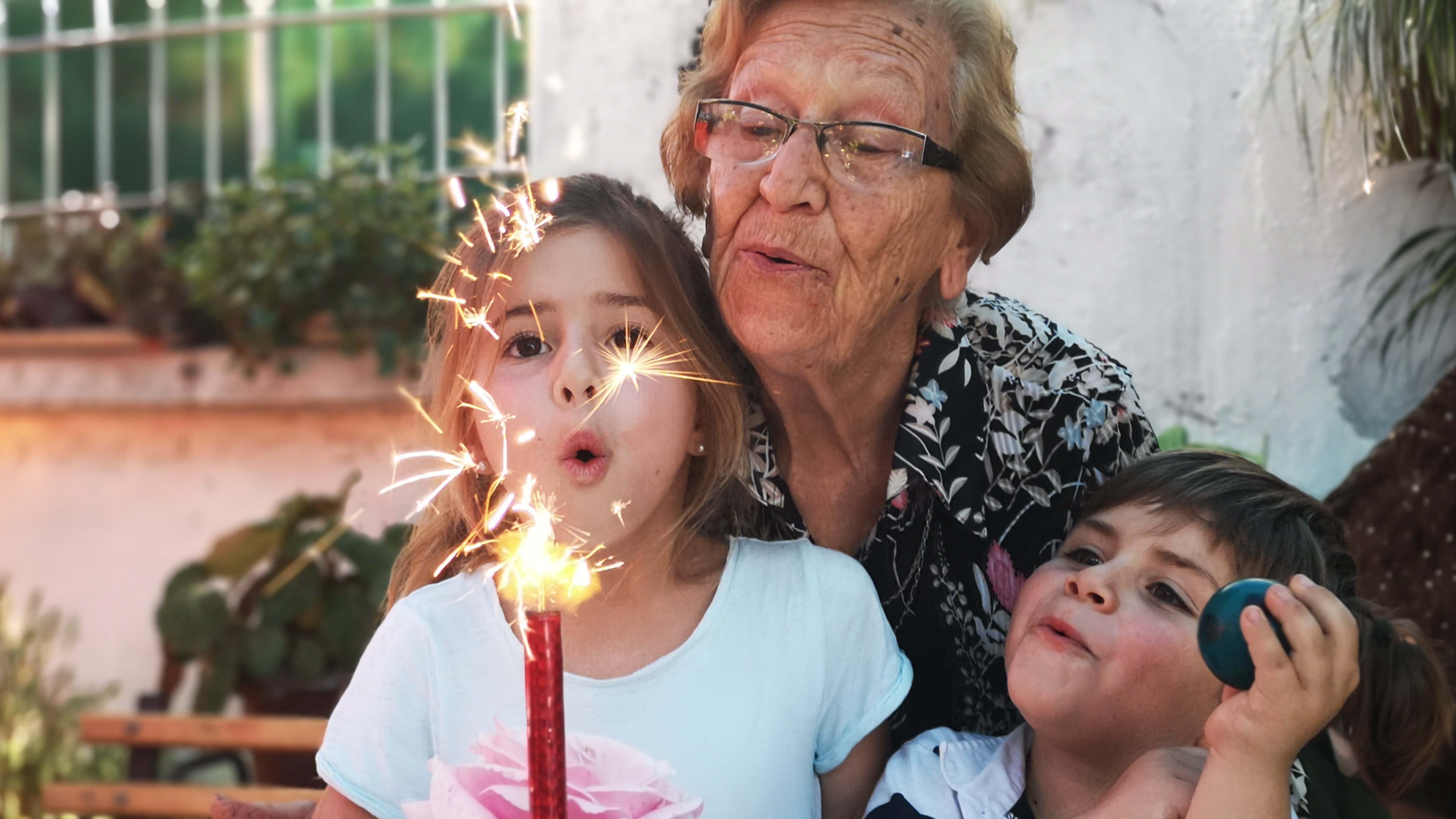 Ältere Person mit Brille und zwei Kindern im Freien; ein Kind hält eine Wunderkerze, während alle drei die Funken aufmerksam betrachten.