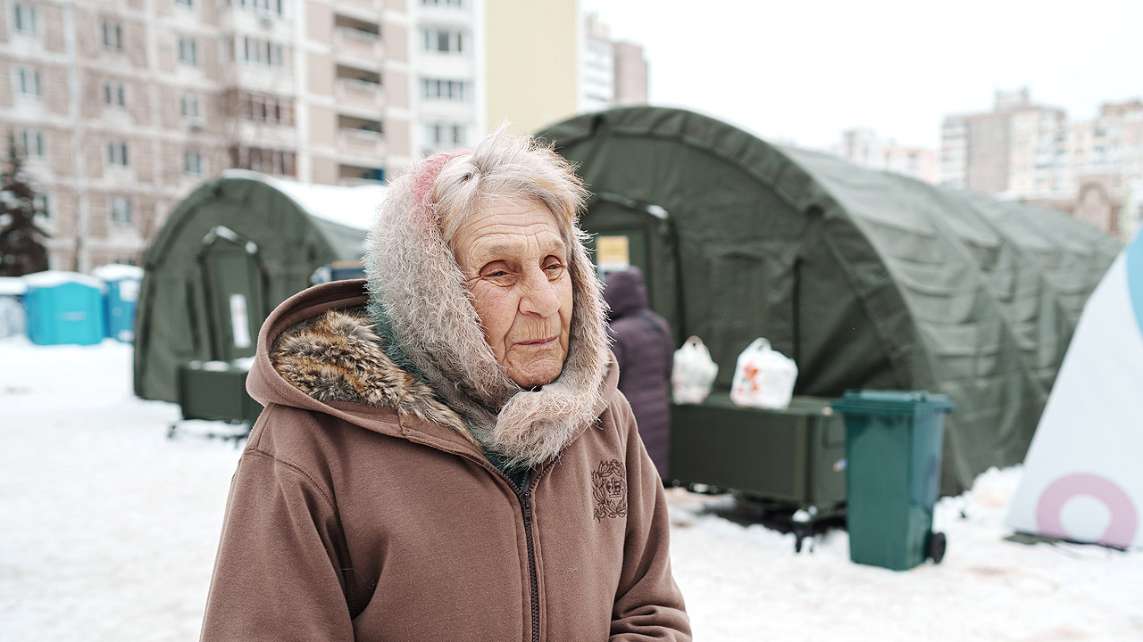 Eine ältere Dame vor einem halbrunden, langen, schwarzen Zelt, alles ist verschneit, dahinter Wohnblocks