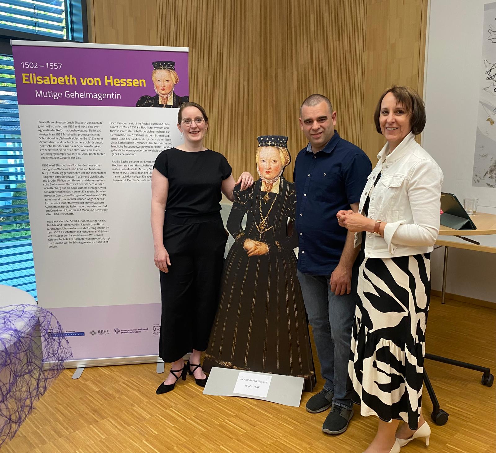 Elisabeth Engler-Starck und Birgit Hamrich gemeinsam mit Martin Röhling im Johanniter-Saal des Dekanats, in ihrer Mitte ein Aufsteller, der Katharina von Bora darstellt, links neben Engler-Starck ein Roll-up mit den wichtigsten Informationen zu Katharina von Bora. Die Menschen stehen nebeneinander und tragen überwiegend dunkle Kleidung.