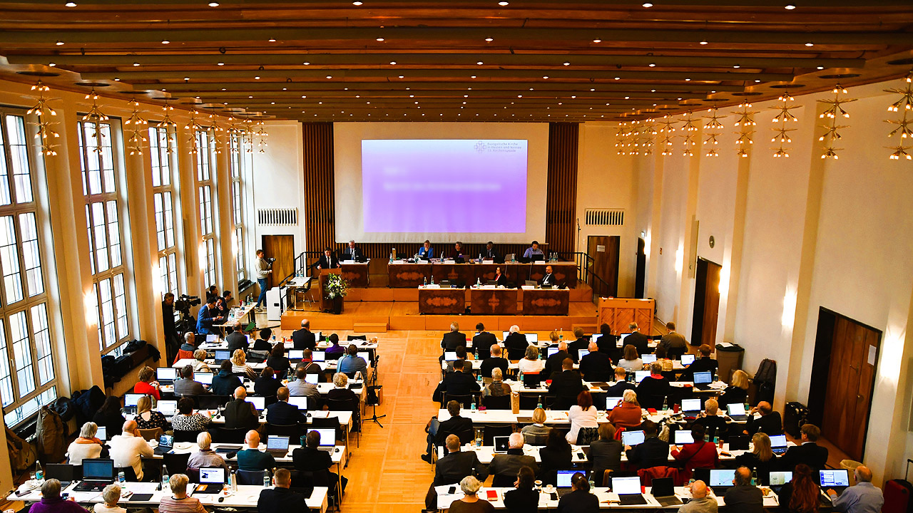 Großer Saal mit Leinwand am Ende, davor viele Bankreihen mit Menschen