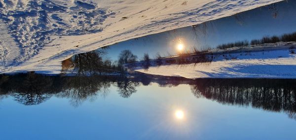 Man sieht einen Fluss in einer Schneelandschaft und die schon untergehende Sonne spiegelt sich im Fluss