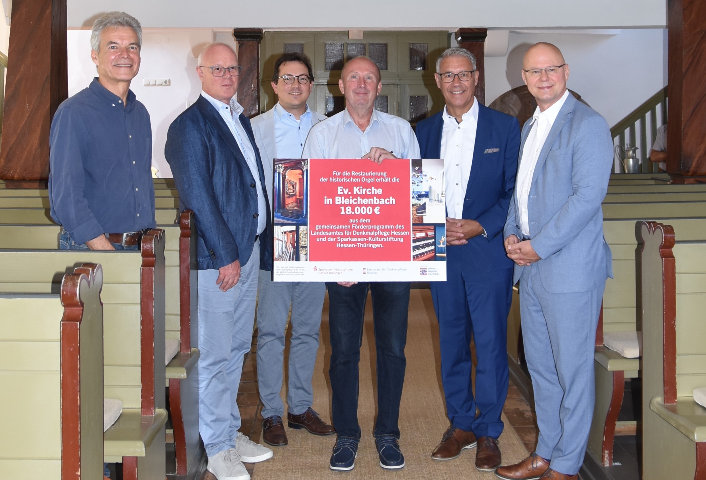 Kirchenvorstansvorsitzender Rolf Appel steht in der Mitte einer Gruppe Männer, , Vertreter von Denkmalpflege und Sparkasse, die meisten in Anzügen, und hält den großen roten, symbolischen Bewilligungsbescheid in die Kamera. Im Hintergrund sieht man die imposante Bleichenbacher Kirchenorgel.