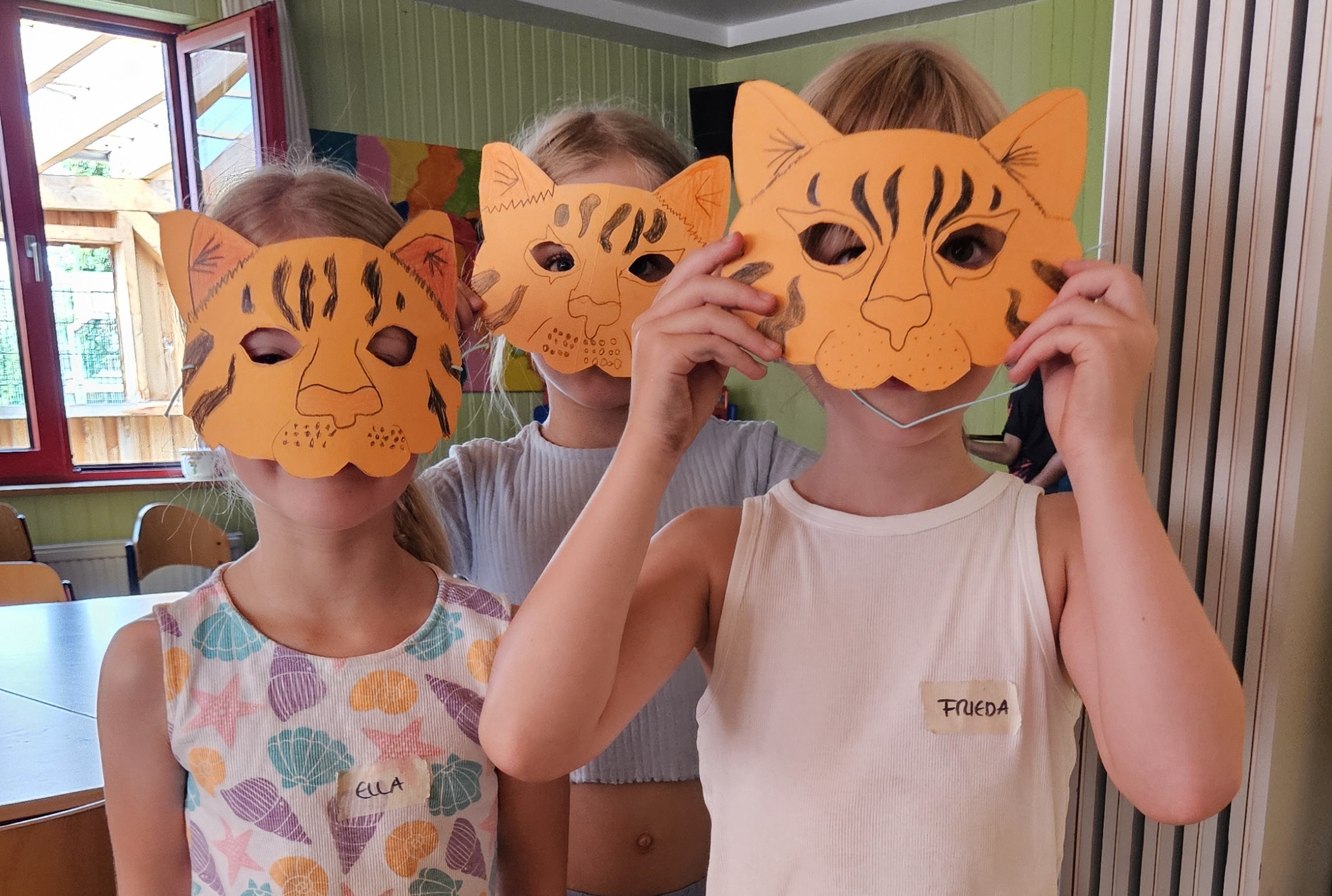 Drei Mädchen halten sich Tigermasken vor das Gesicht, die selbst aus Pappe gebastelt und mit Buntstiften bemalt haben. 