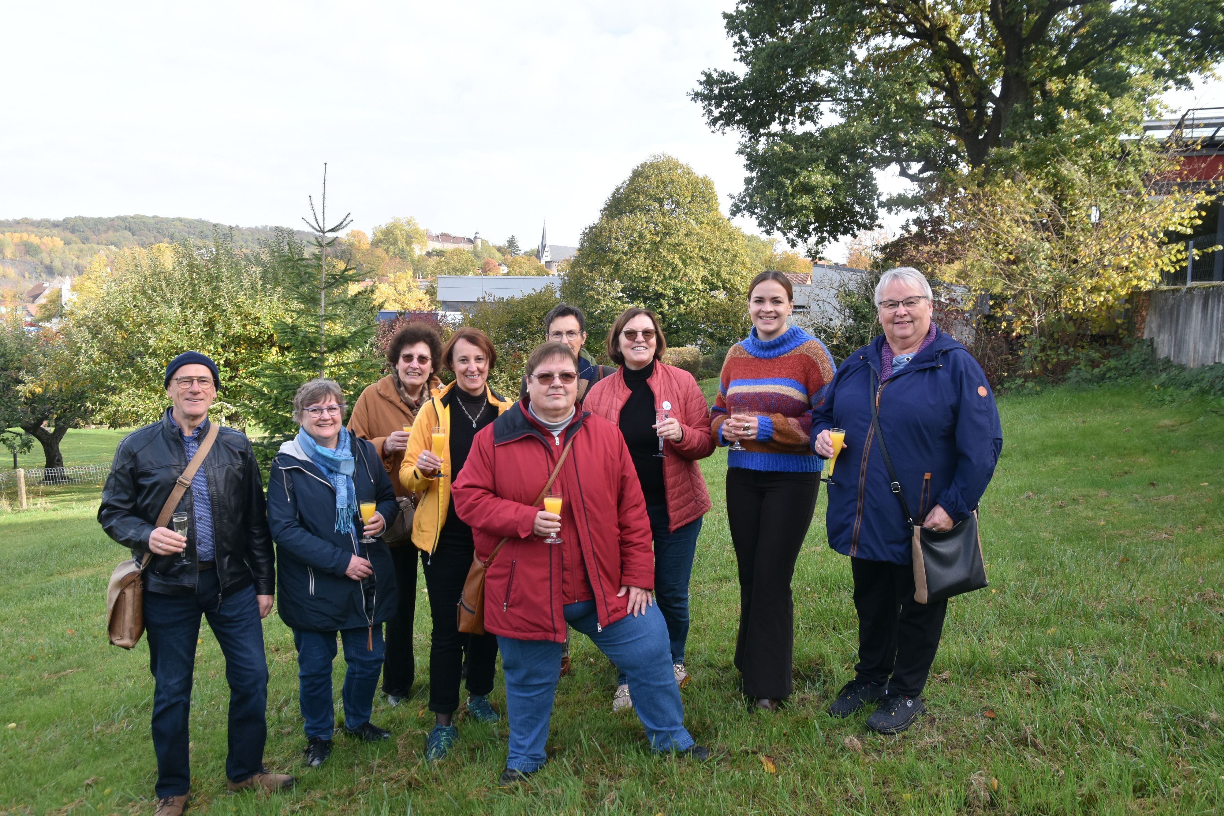 Eine neunköpfige Gruppe, acht Frauen, ein Mann, stehen in einer Wiese vor Obstbäumen und halten Sektgläser in der Hand. Alle tragen warme Kleidung. Im Hintergrund sieht man das Ortenberger schloss und den Turm der Marienkirche.