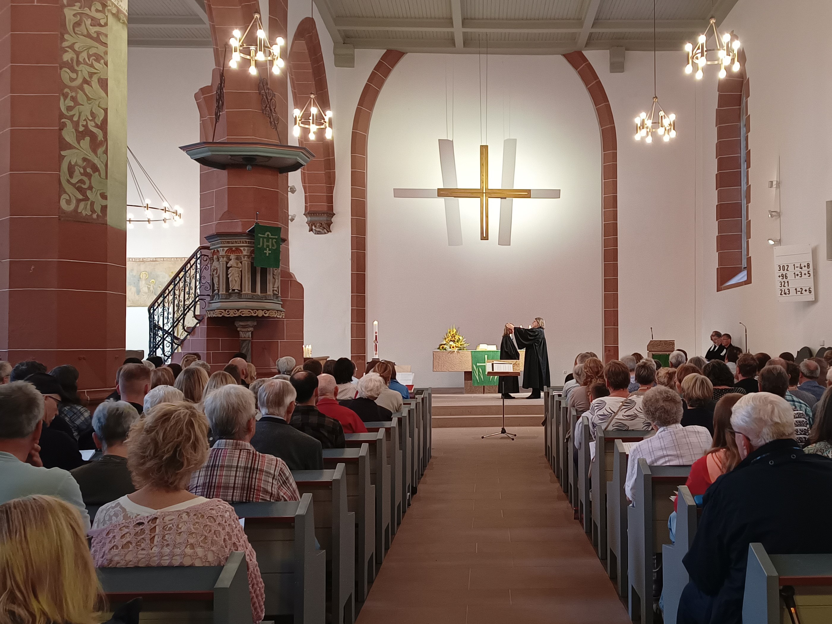 Blick in eine vollbesetzte Kirche. Menschen sitzen in den Bankreihen. Vor dem Altar stehen vorne zwei Pfarrerinnen.