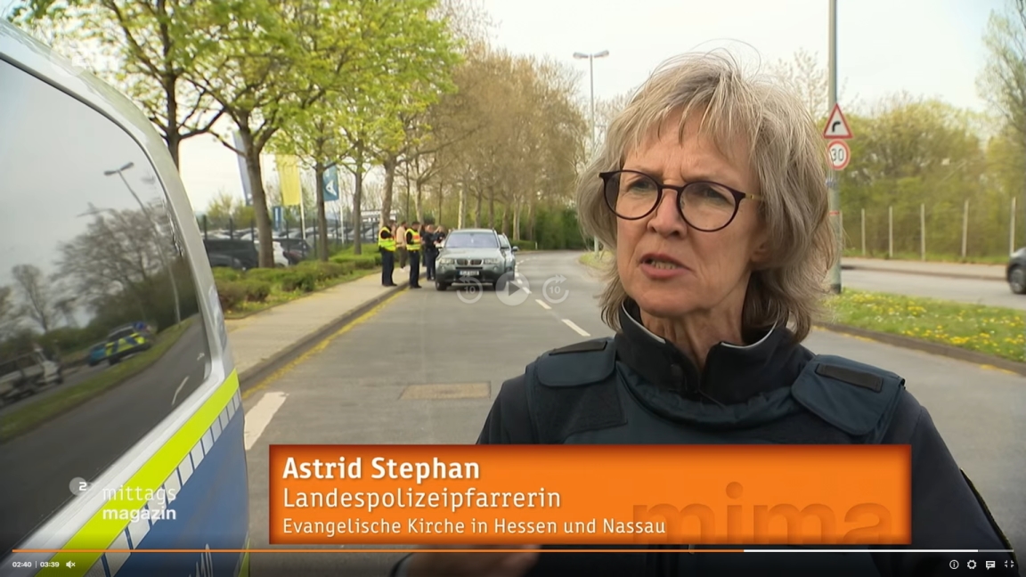 Zu sehen ist ein Screenshot des Fernsehbeitrag mit der Pfarrerin auf einer Straße neben einem Polizeifahrzeug.