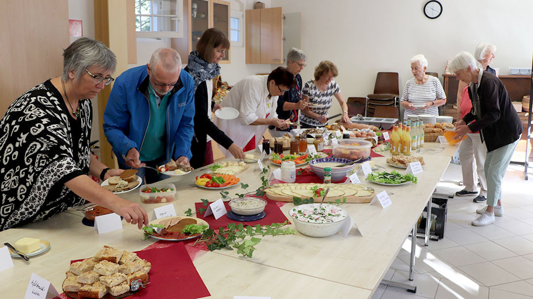 Mehrere Menschen bedienen sich gemeinsam an einem reich gedeckten Frühstücksbuffet mit Brot, Aufstrichen, Gemüse und Getränken.