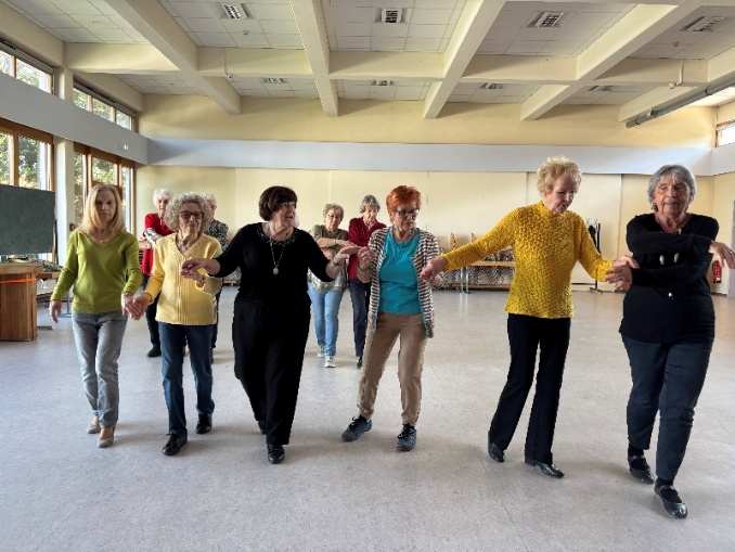 Eine Gruppe älterer Menschen tanzt gemeinsam in einem hellen, großen Saal und hält sich dabei an den Händen. Die Personen bewegen sich in einer Reihe auf die Kamera zu, wirken konzentriert und fröhlich, während durch die großen Fenster Tageslicht in den Raum fällt.