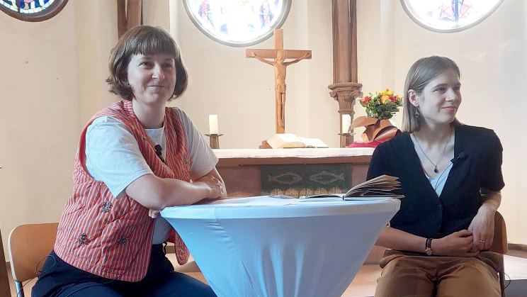 Zwei Frauen sitzen an einem Stehtisch vor dem Altar einer Kirche; auf dem Tisch liegt ein aufgeschlagenes Buch, im Hintergrund sind ein Kreuz und Blumen zu sehen.