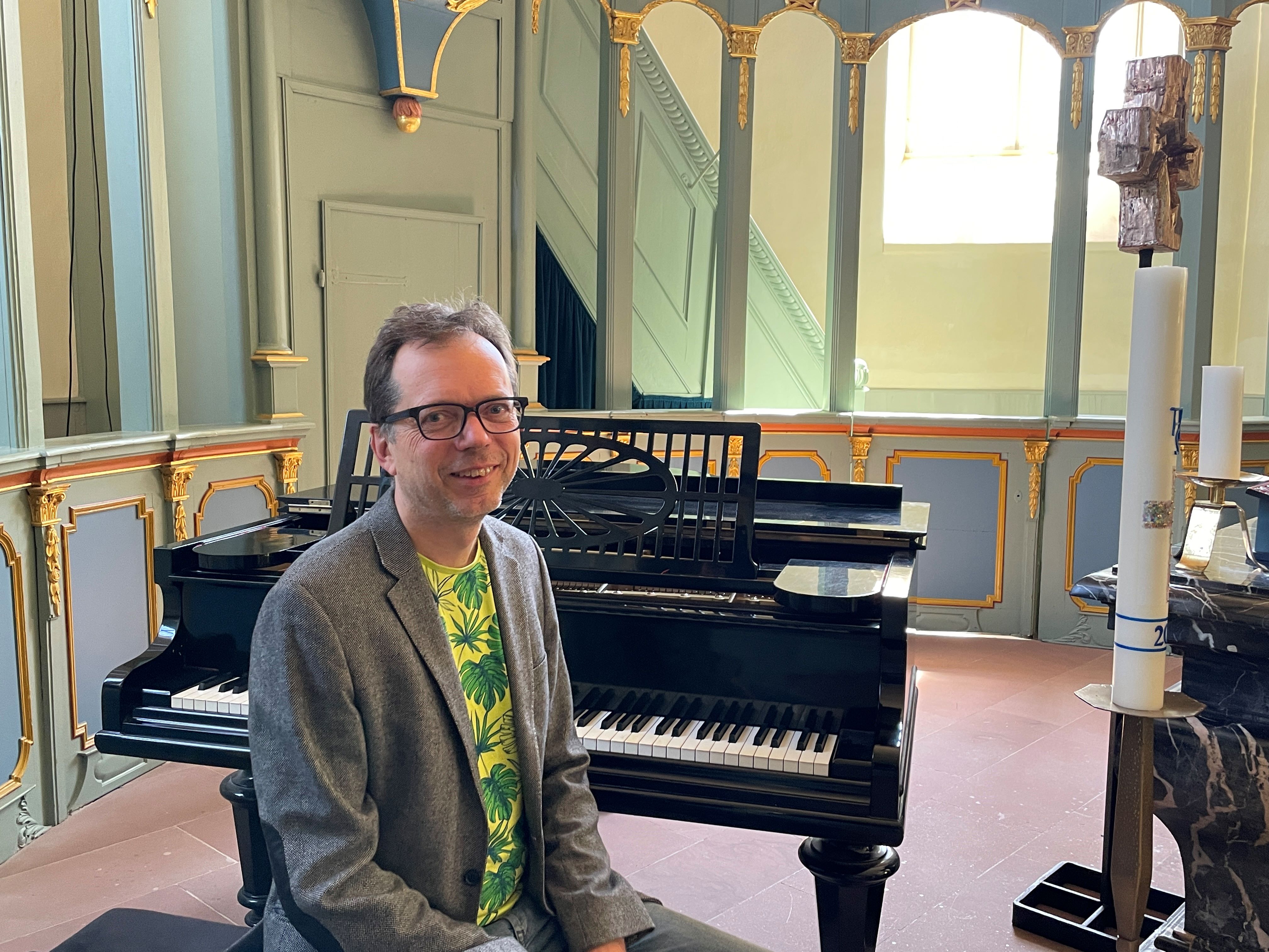 Dekanatskantor Thomas Wächter sitzt an einem Flügel im Altarraum der Evangelischen Kirche Wehen. Dieser wird vom hellen Sonennlicht, dass durch die Fenster kommt, erhellt.