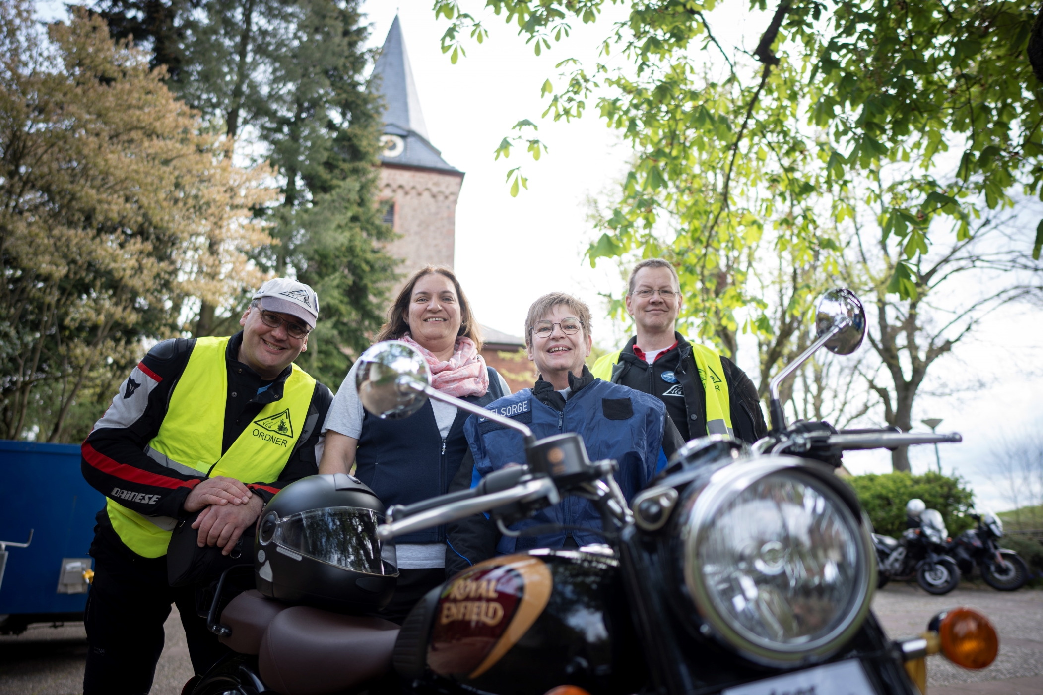 Die vier Personen stehen hinter einem Motorrad. Im Hintergrund ist die Kirche zu sehen.