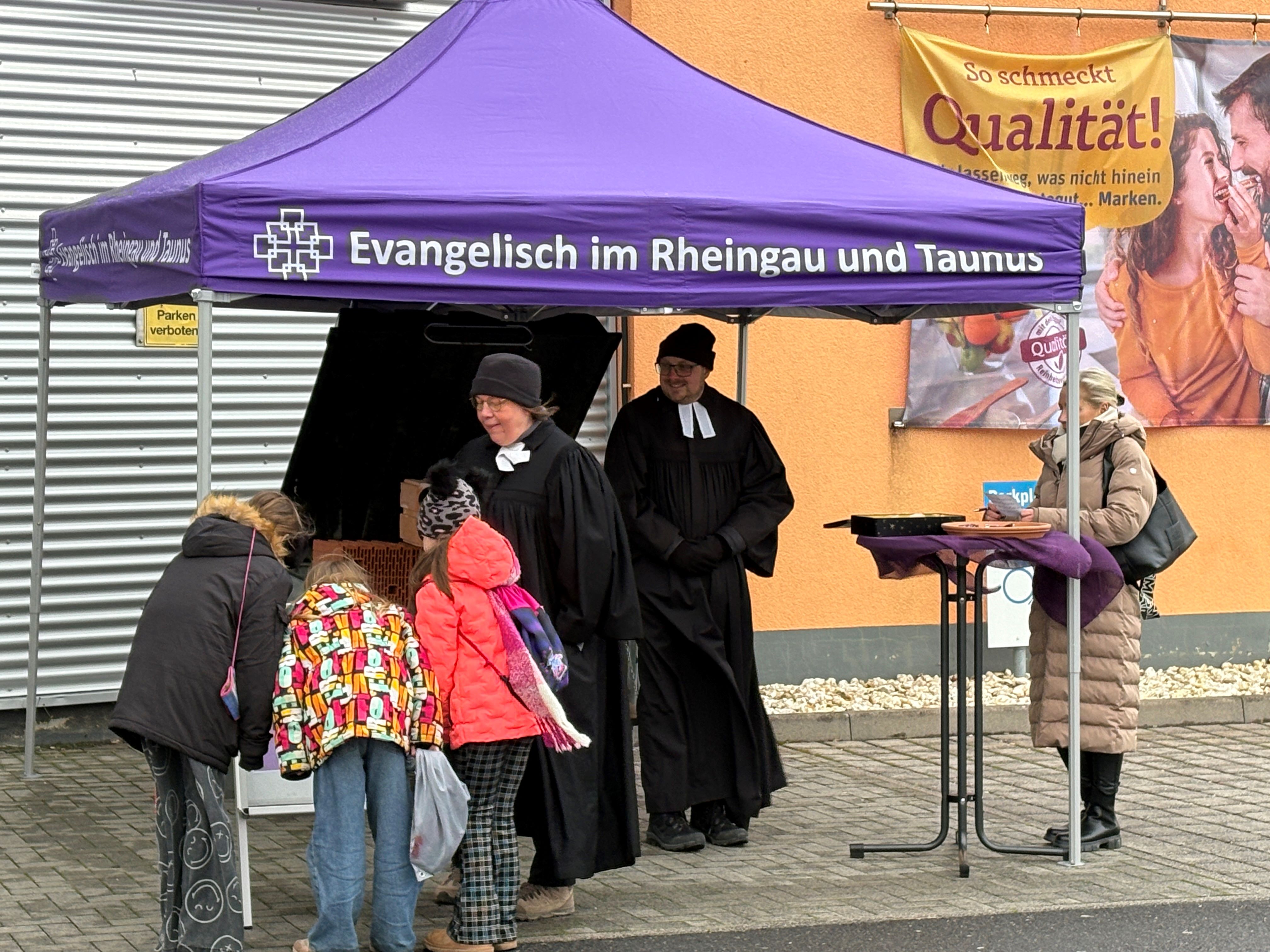 Unter einem lila Pavillon steehen eine Pfarrerin und ein Pfarrer im Talar und laden Menschen an einem Stehtisch ein, Karten auszufüllen. Zur Aktion "Verzeih' mir!" Segensaktion am Busstag