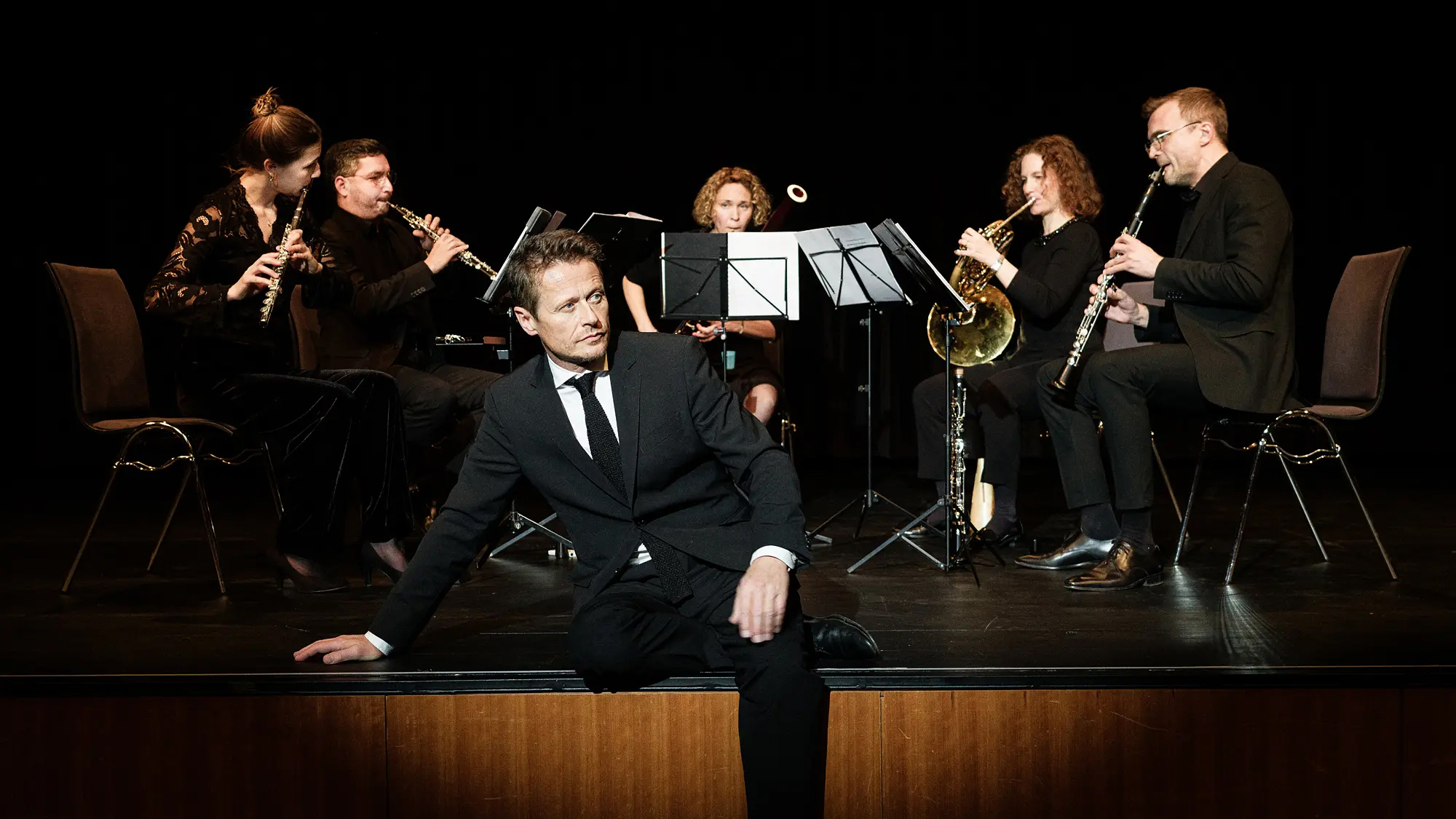 Ein Mann im schwarzen Anzug sitzt nachdenklich am vorderen Rand einer Bühne. Hinter ihm musiziert ein fünfköpfiges Bläserensemble in schwarzer Kleidung mit Flöte, Oboe, Fagott, Horn und Klarinette vor dunklem Hintergrund.