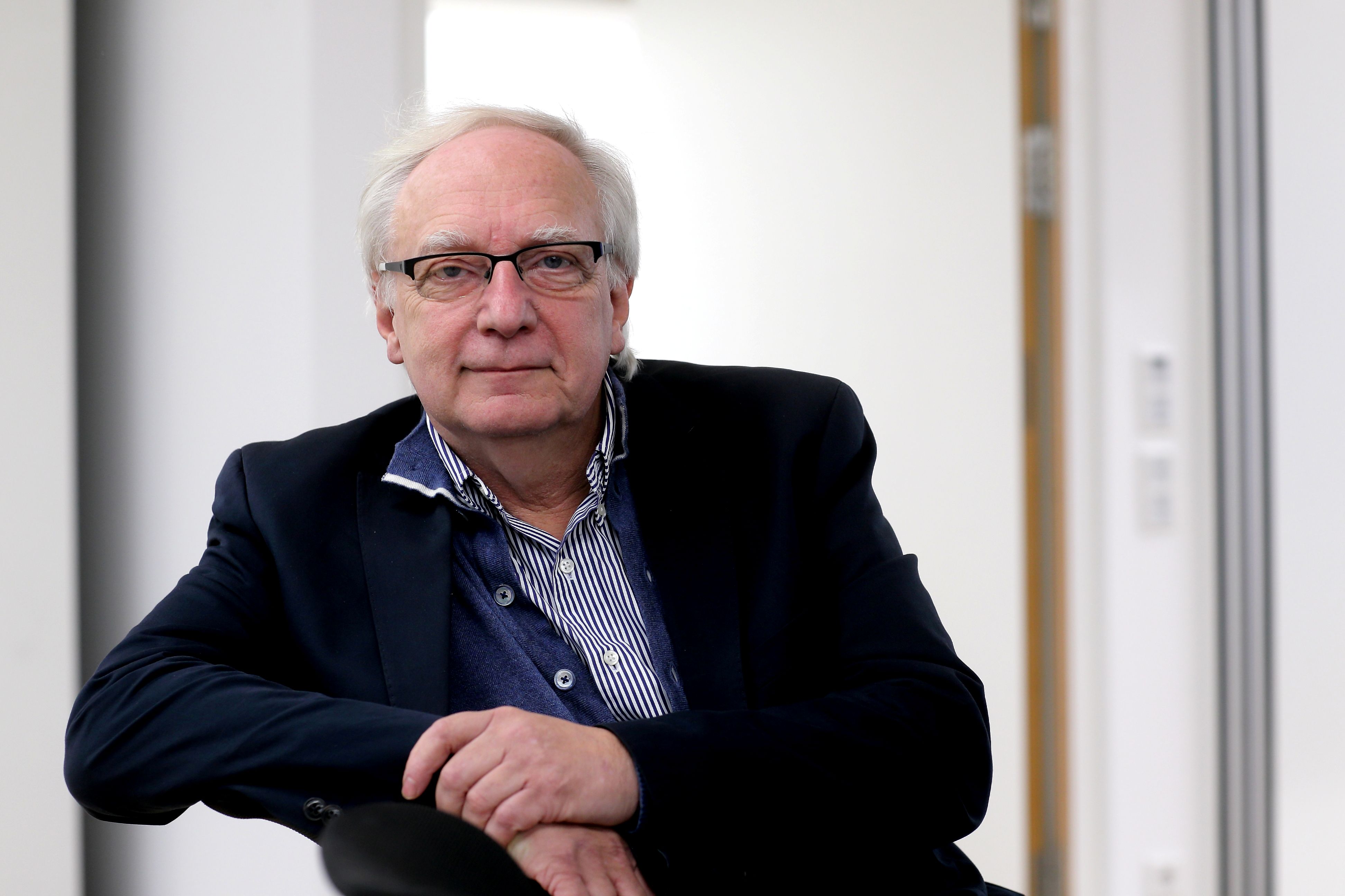 Claus Leggewie, schütteres graues Haar, Brille, Hemd und dunkles Sakko, sitzt zur Seite gedreht auf einem Stuhl, den rechten Arm über die Lehne gelegt, die linke Hand liegt über der rechten. Er schaut aufmerksam in die Kamera.
