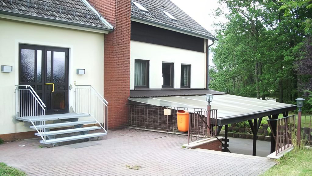 Eingangsbereich des Gerhart-Hauptmann-Hauses in Scharbach. Links führt eine Metalltreppe mit Geländer zu einer dunklen Doppeltür. Rechts schließt sich eine überdachte Zufahrt an. Das Gebäude mit heller Fassade und rotem Backsteinschornstein ist von Bäumen umgeben.
