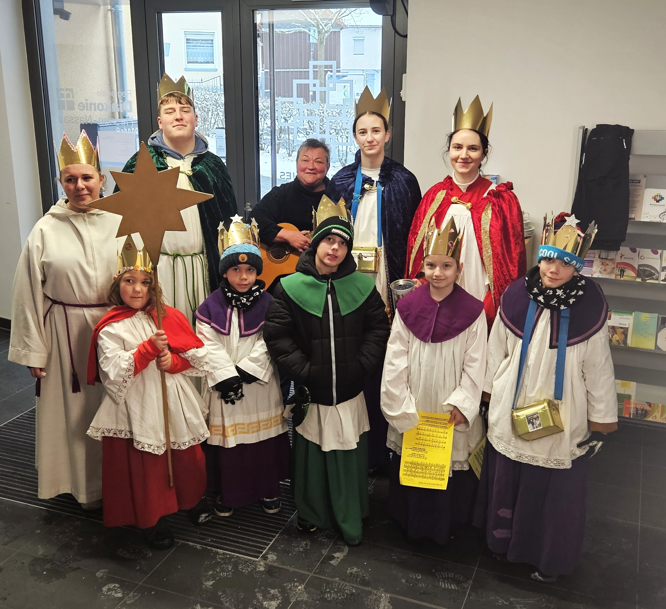 Neun Kinder und Jugendliche und eine Begleitperson stehen im Foyer des Margaretha-Pistorius-Hauses. Sie sind verkleidet als Sternsinger und tragen Messdiener-Gewänder, goldene Kronen und führen einen goldenen Stern mit sich.