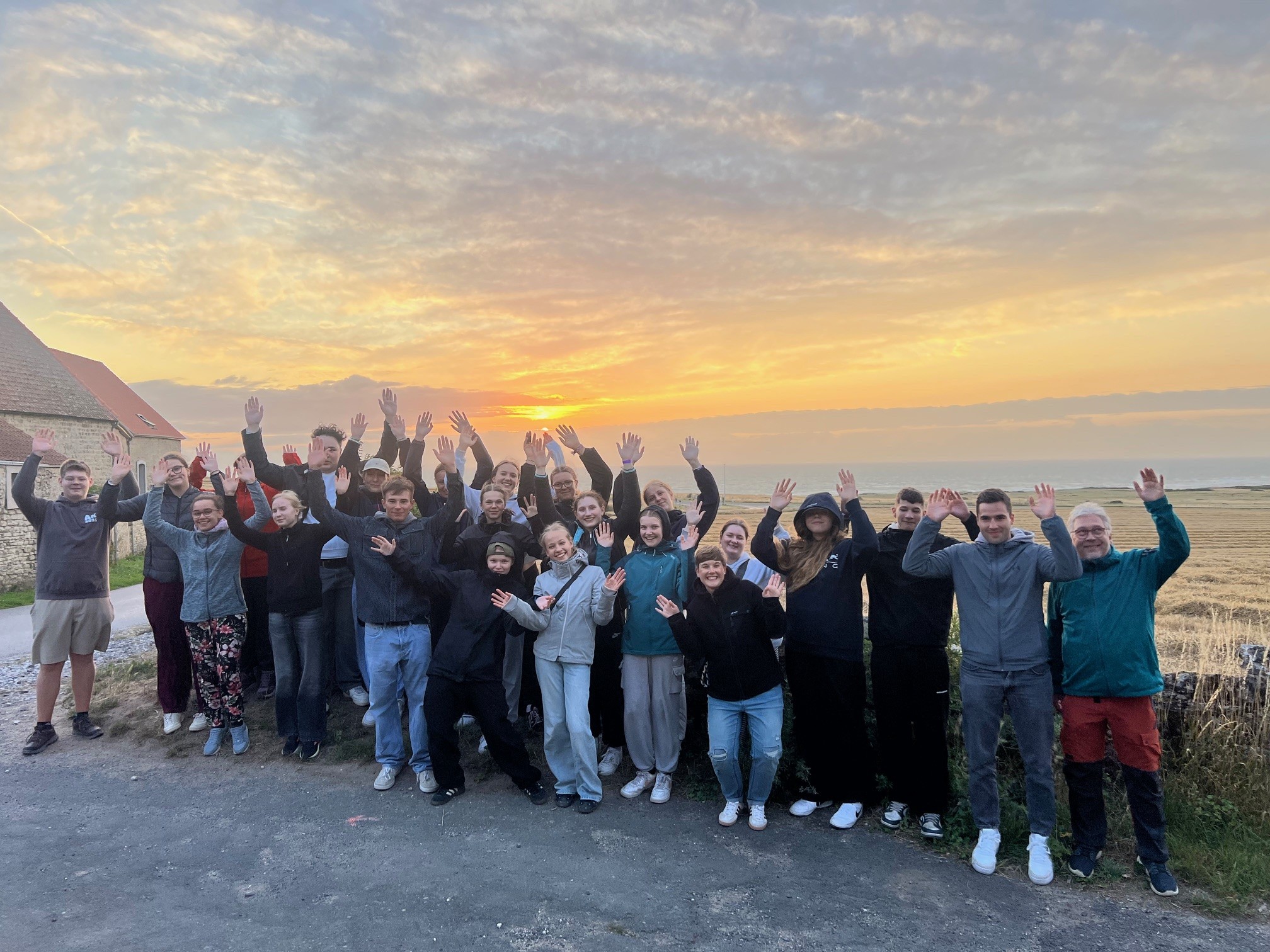 Eine Gruppe junger Menschen steht an einem Weg, alle winken in die Kamera. Im Hintergrund Gräser, Sand und weiter Weg das Meer. Darüber ein Himmel in Orange-und Grautönen.
