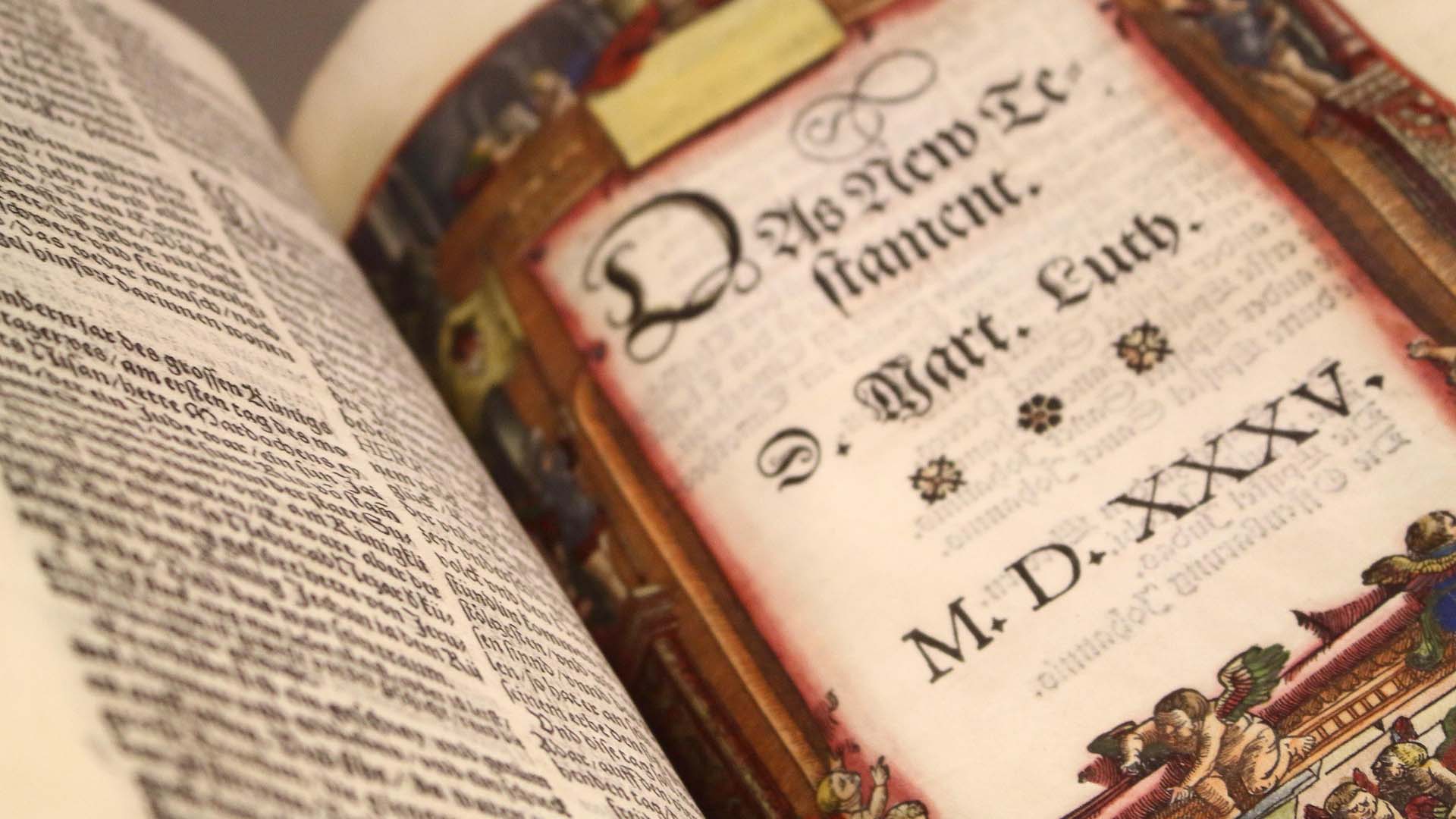 Nahaufnahme einer aufgeschlagenen historischen Lutherbibel. Rechts ist das kunstvoll gestaltete Titelblatt des Neuen Testaments mit alter deutscher Schrift, farbigen Verzierungen und der Jahreszahl 1535 zu sehen. Links befindet sich eine dicht bedruckte Buchseite.