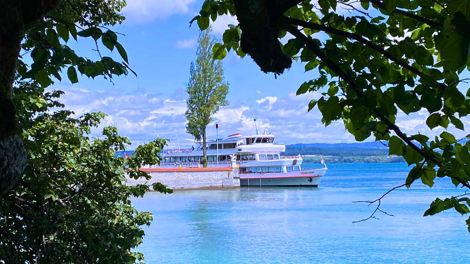 Ein Fahrgastschiff liegt an einem Steg auf einem See, eingerahmt von dichtem Laub und Bäumen, unter einem teilweise bewölkten Himmel.