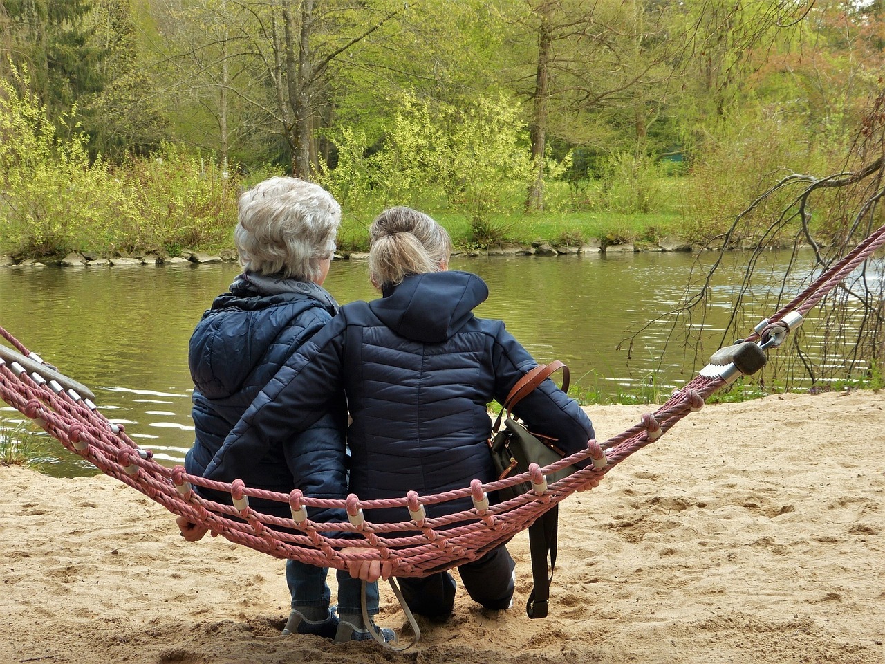 Zwei Frauen sitzen mit dem Rücken zur Kamera in einer Hängematte am Ufer eines Gewässers