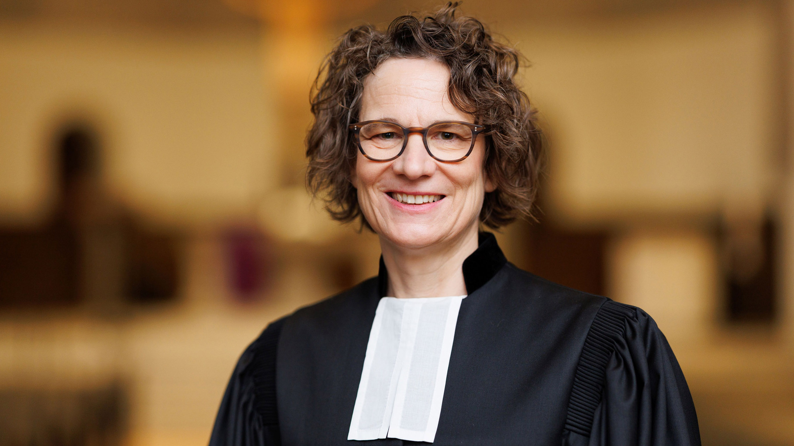 tietz_christiane_talar_da25_q_peter_bongard_2700_16zu9.jpg Christiane Tietz, Kirchenpräsidentin der Evangelischen Kirche in Hessen und Nassau (EKHN), lächelt in die Kamera. Sie trägt eine Brille und einen schwarzen Talar mit weißem Beffchen. Der Hintergrund ist unscharf und in warmen Farbtönen gehalten.
