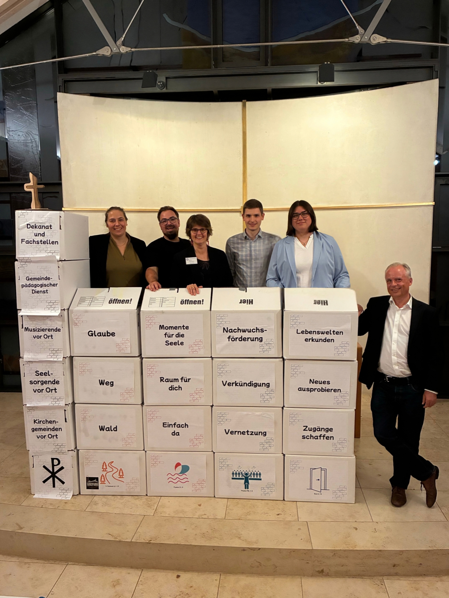 Eine Kirche aus Umzugskartons gebaut, mit Wörtern zu den Arbeitsbereichen der vier neuen Kolleginnen und Kollegen. Über die Kirche schauen die Mitarbeiter, daneben steht Präses André Jacob