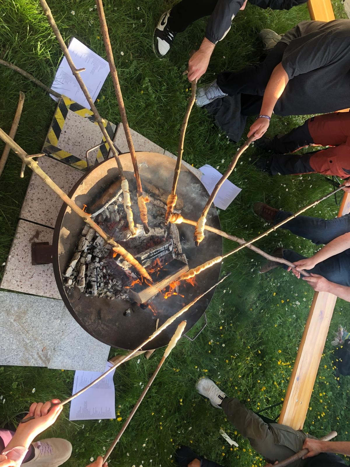 Hier ist eine einfache Beschreibung des Bildes:  Das Bild zeigt eine Gruppe von Menschen, die um ein Lagerfeuer stehen und Stockbrot backen. Sie halten lange Stöcke mit Teig über die Flammen. Um das Feuer herum liegt Gras und es sind einige Blätter Papier zu sehen. Im Hintergrund sitzen einige Leute auf einer Bank.