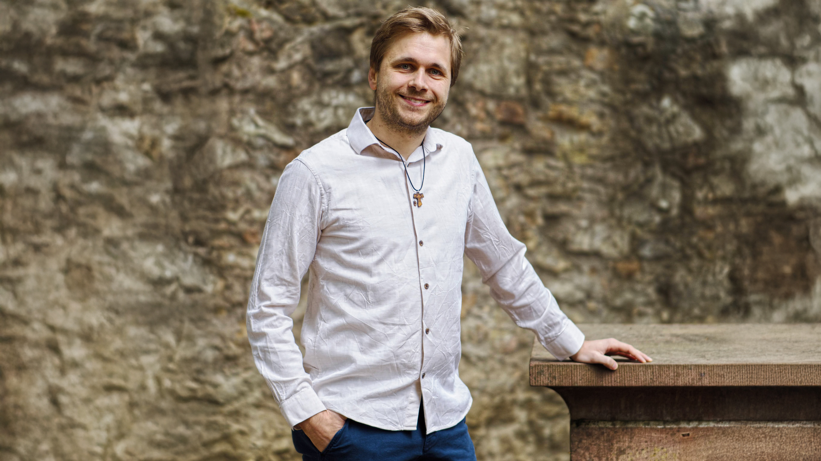 Joshua van der Linden steht lächelnd vor einer alten Natursteinmauer. Er trägt ein helles Hemd, eine dunkelblaue Hose und eine Kreuzkette. Eine Hand steckt in seiner Hosentasche, die andere ruht auf einer steinernen Brüstung.