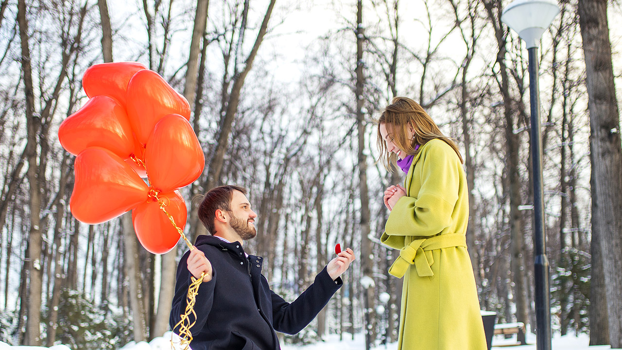 Ein Mann geht vor einer Frau in die Knie, er hat viele rote Herz-Luftballons dabei, es ist draußen im Schnee.