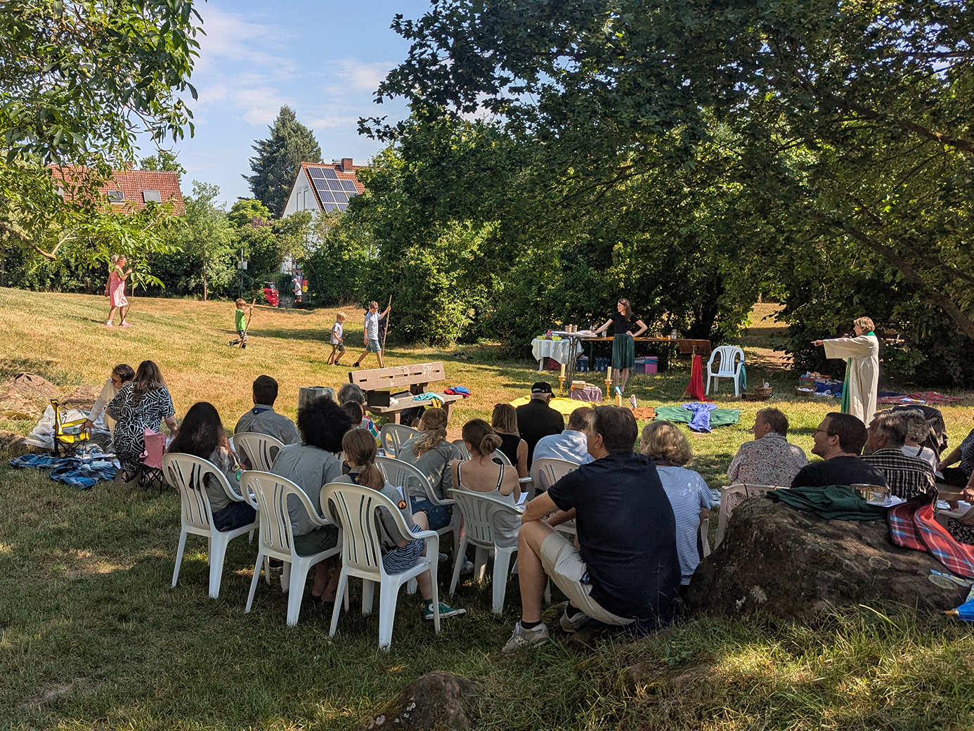 ein Gruppe von Menschen sitzt unter einem Baum und hört einer Pfarrerin zu. Kinder spielen