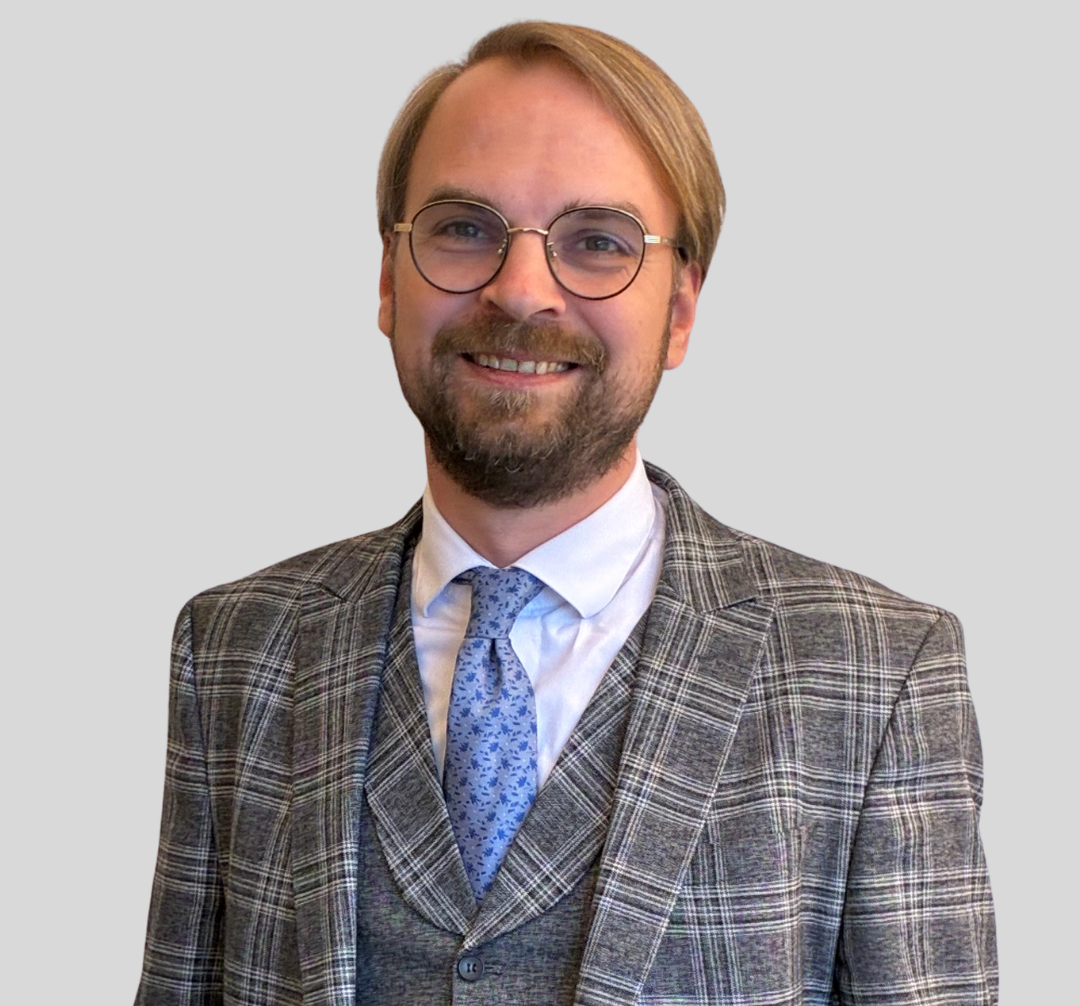 Georg Bloch-Jessen trägt einen karierten Anzug mit Weste und eine blaue Krawatte, seine Haare sind blond. Er trägt eine Brille.