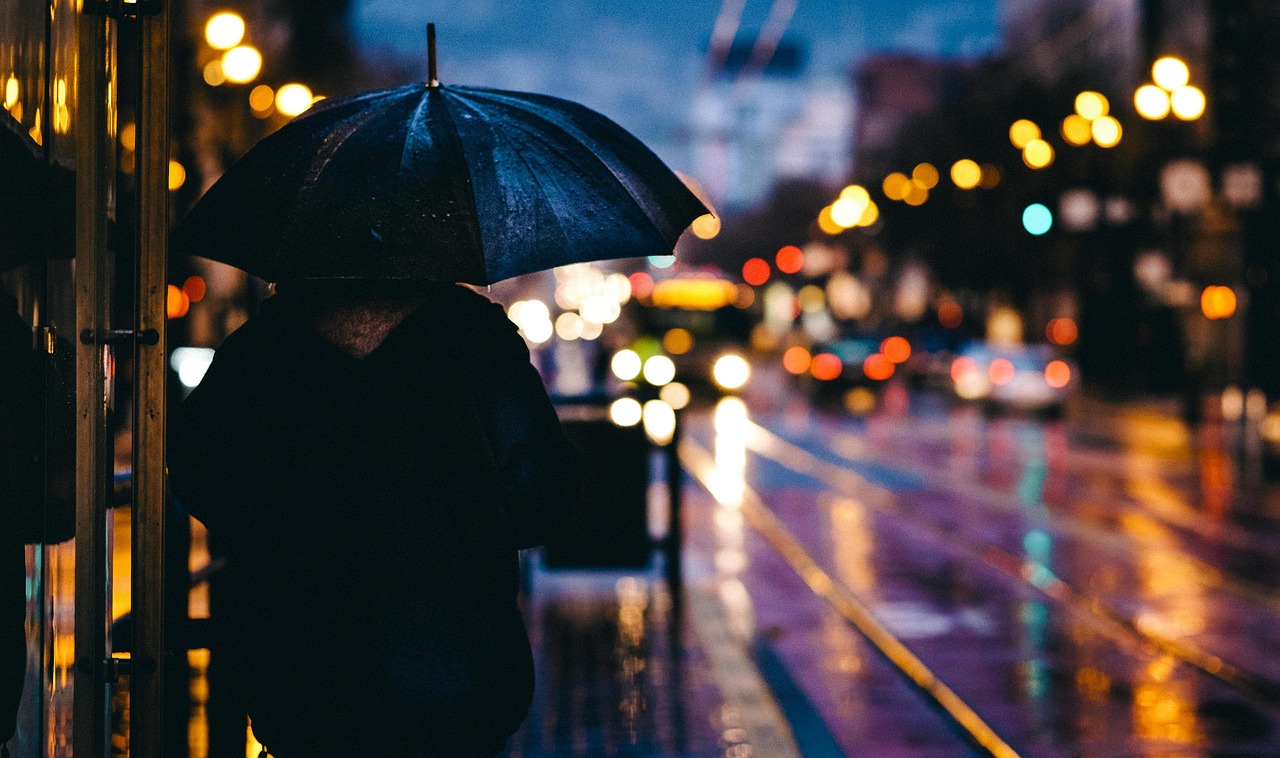 „Eine Person mit Regenschirm steht bei Regen an einer beleuchteten Straße in der Dämmerung. Das unscharfe Stadtlicht erzeugt eine nachdenkliche und ruhige Atmosphäre.“