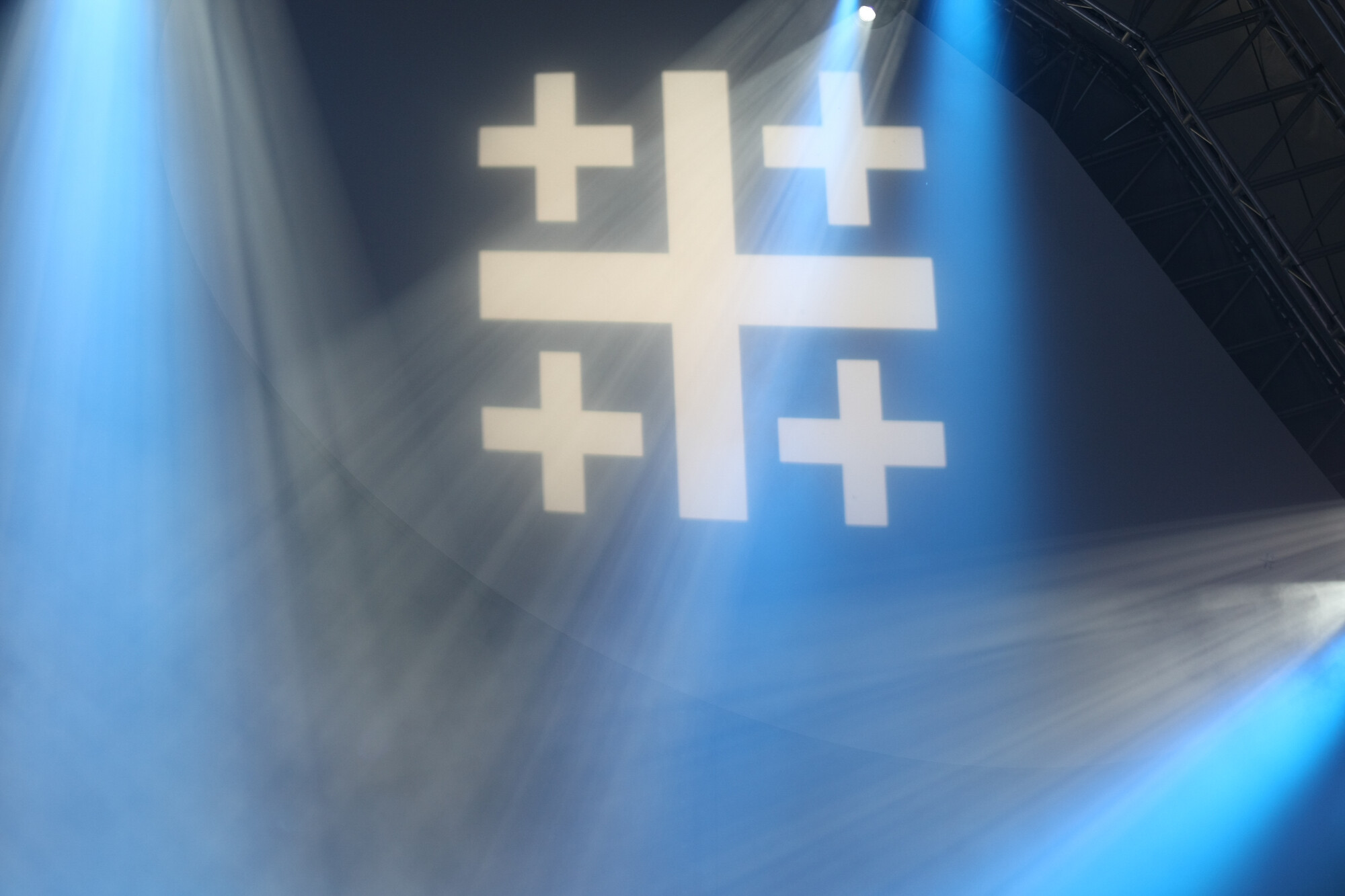 Ein Jerusalemkreuz wird in einer blauen Halle projiziert und Scheinwerfer bestrahlen es